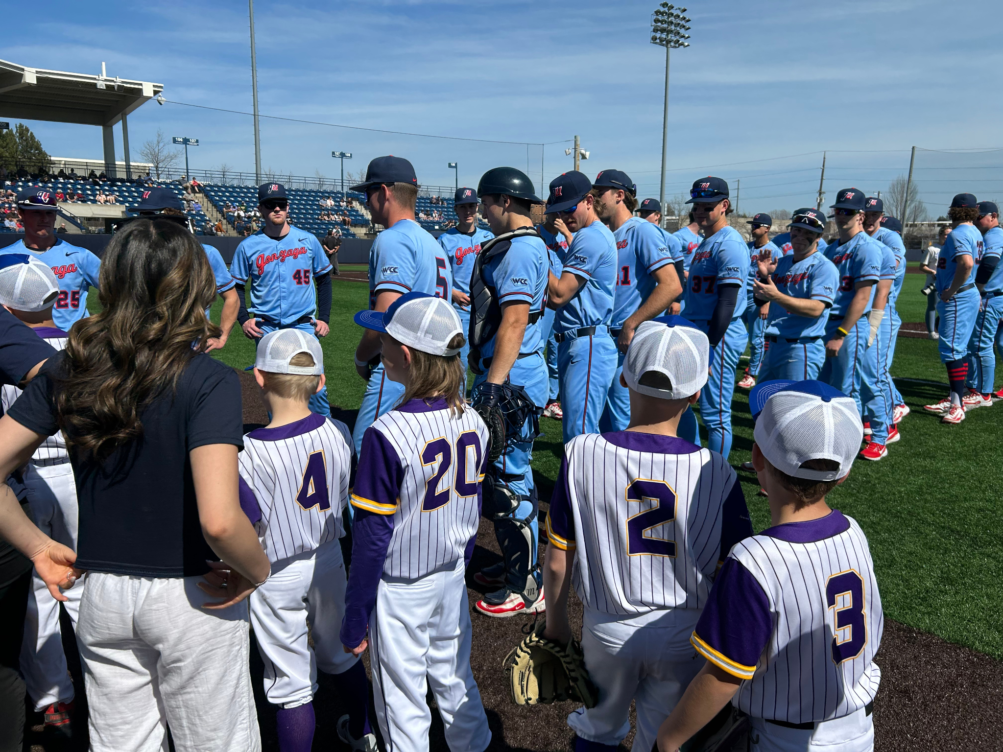 A group of children and adults, wearing baseball uniforms, gathered on a baseball field. The children are in white and purple jerseys with numbers on the back, while the adults wear blue uniforms. They appear to be having a team meeting or interactin