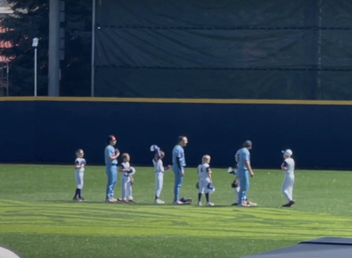 Kids and adults in baseball uniforms standing on a field during a ceremony or national anthem.