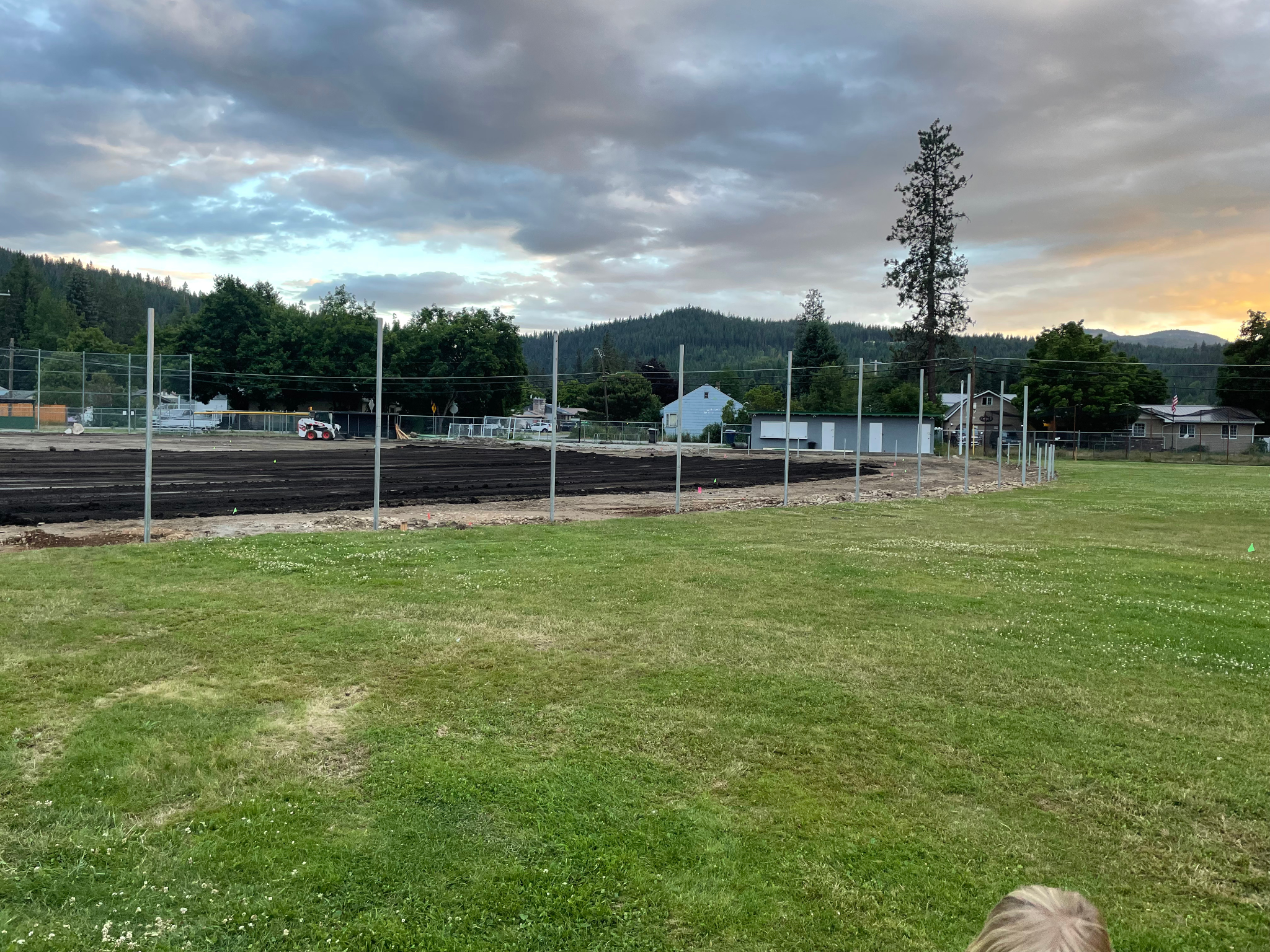 Outdoor field with construction equipment, surrounded by partial fencing, grassy area, trees, houses, and mountain backdrop under a cloudy sky.
