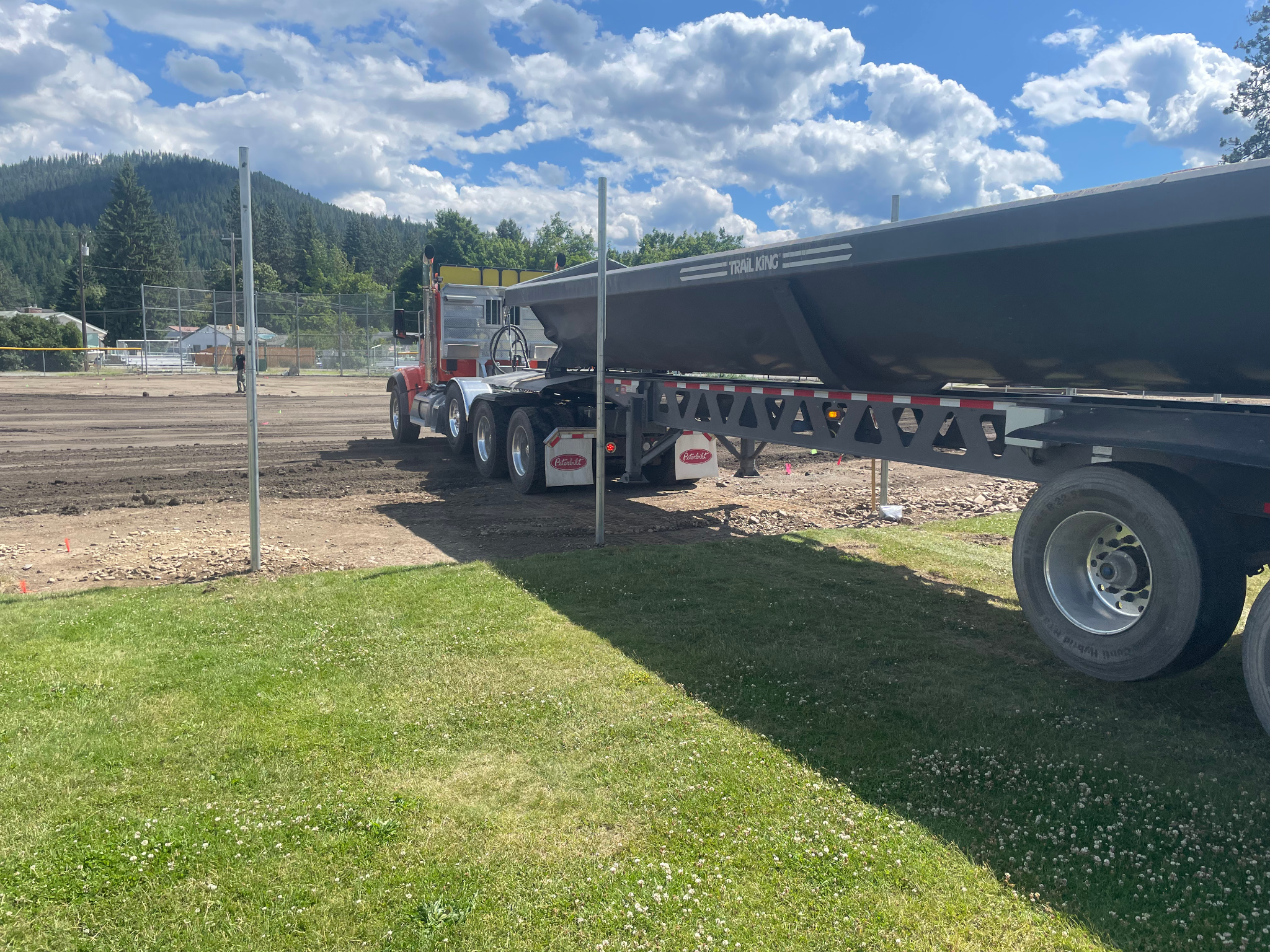 Large flatbed truck parked on a grassy area next to a dirt field, with trees and cloudy sky in the background.