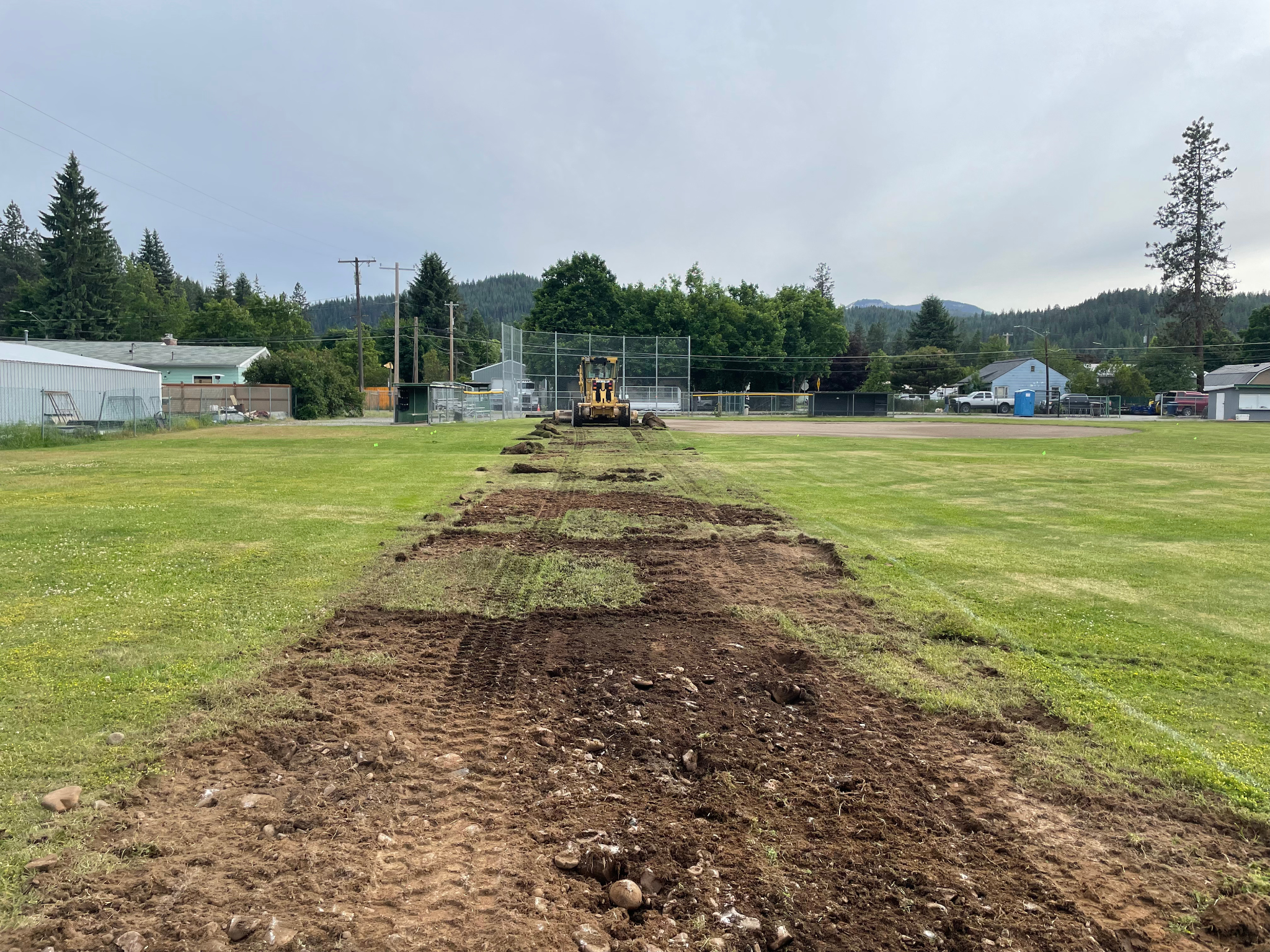 A construction vehicle working on a grass field, with freshly dug earth and a backdrop of trees and buildings.