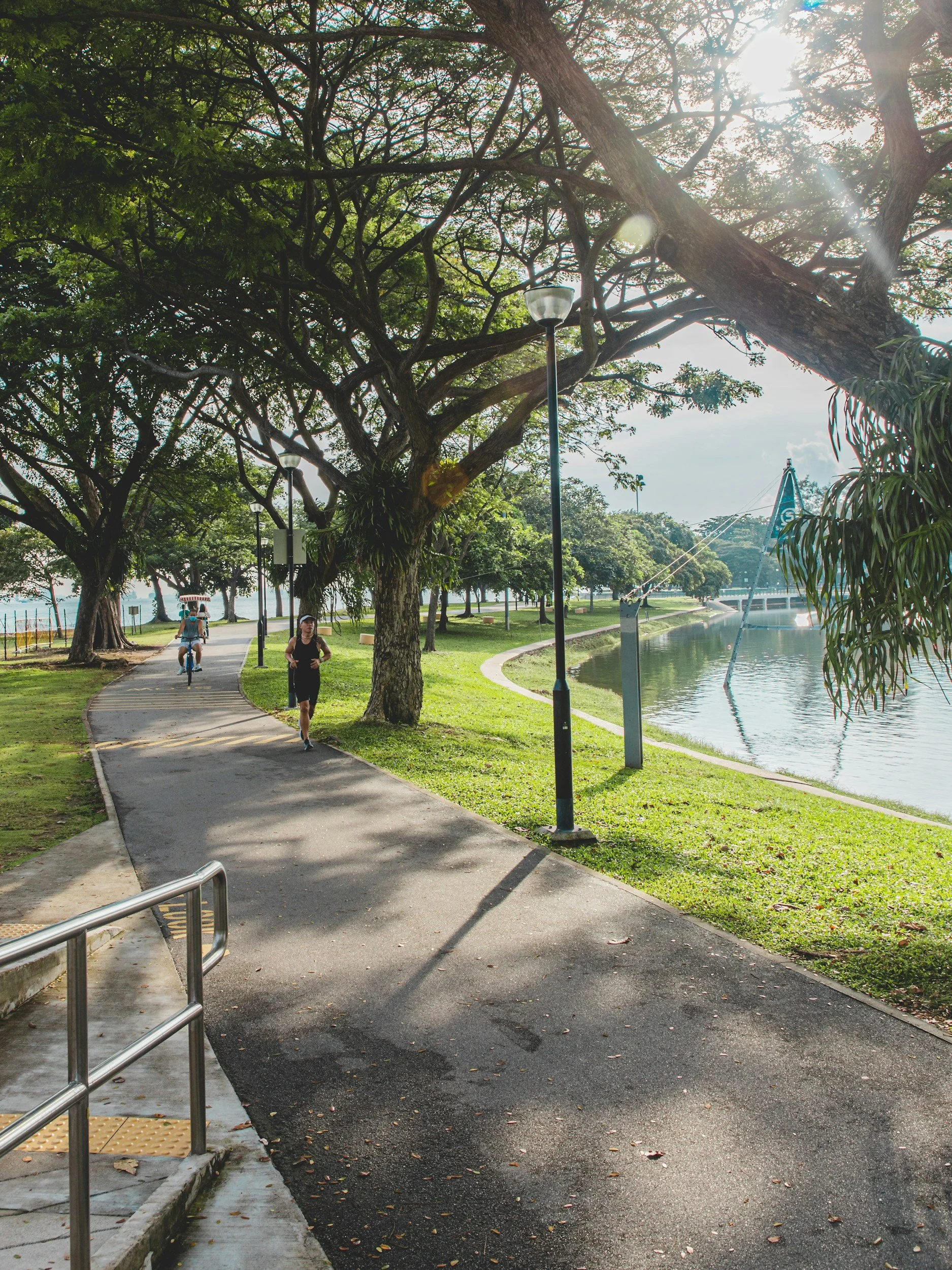 A park pathway with trees and grass on either side, and a waterway to the right. Two people are walking and a cyclist is riding on the pathway. There are streetlights along the pathway, and a bridge is visible over the waterway in the background. The scene is illuminated by sunlight.