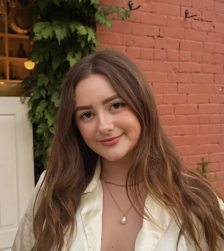 A young woman with long, wavy brown hair smiling at the camera, standing outdoors in front of a red brick wall and green leafy plants.