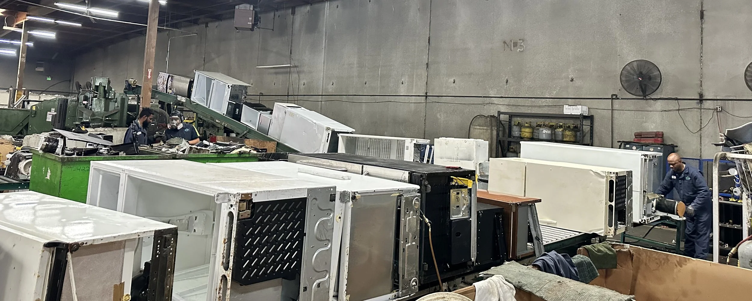 Workers in a warehouse operating and repairing industrial equipment and machinery among disassembled appliances, with a concrete wall, shelves, and ventilation fans visible in the background for recycling