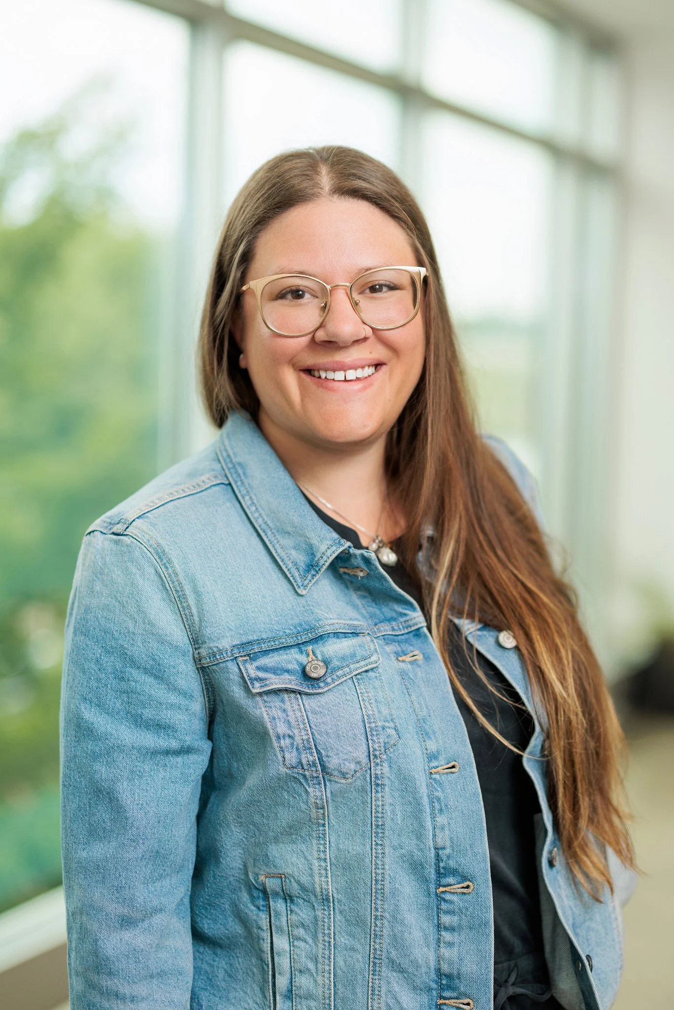 A woman with long brown hair wearing glasses and a denim jacket, smiling indoors near large windows.