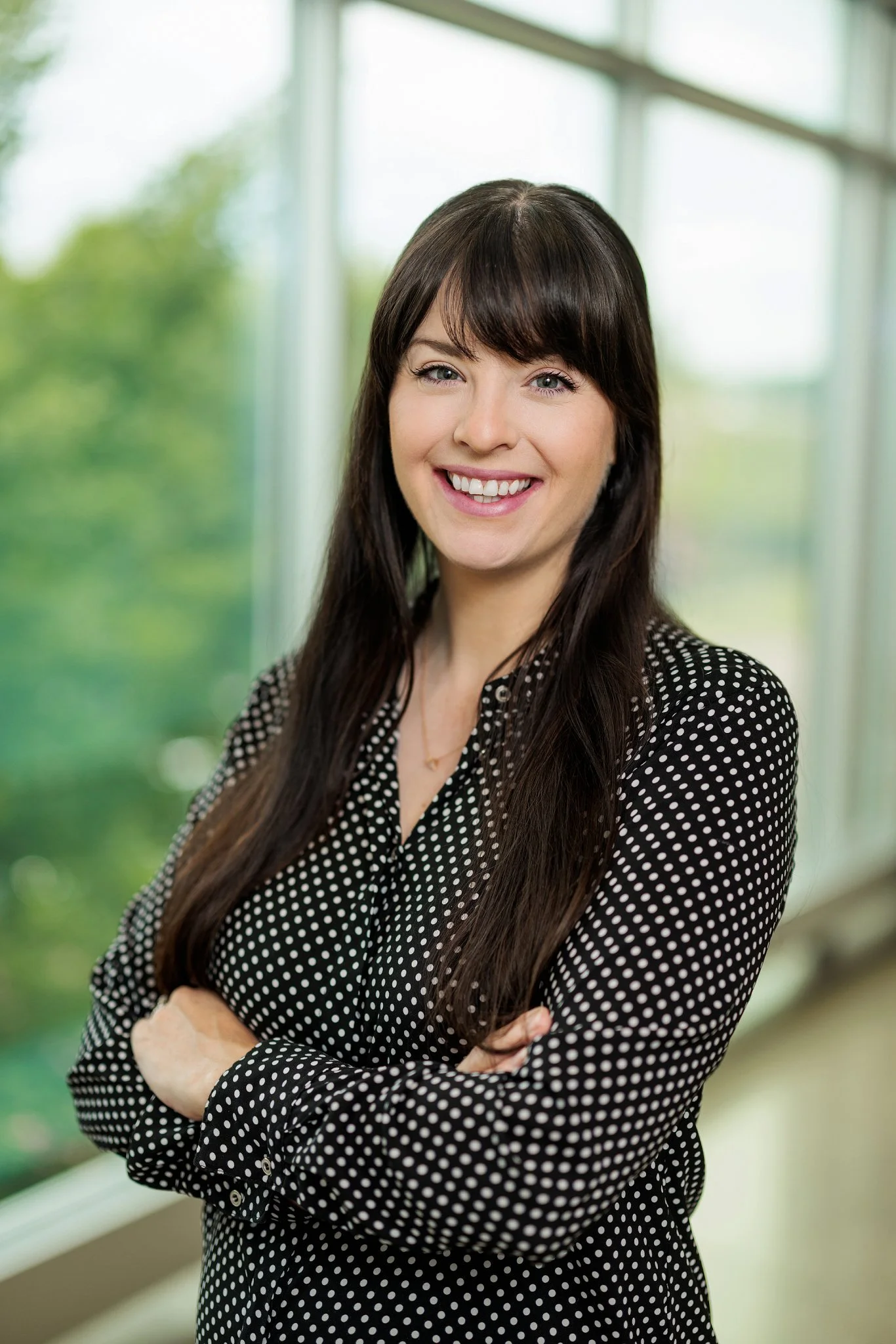A woman with long brown hair and a fringe smiling, wearing a black dress with white polka dots, standing with arms crossed in front of large windows with a green outdoor view.