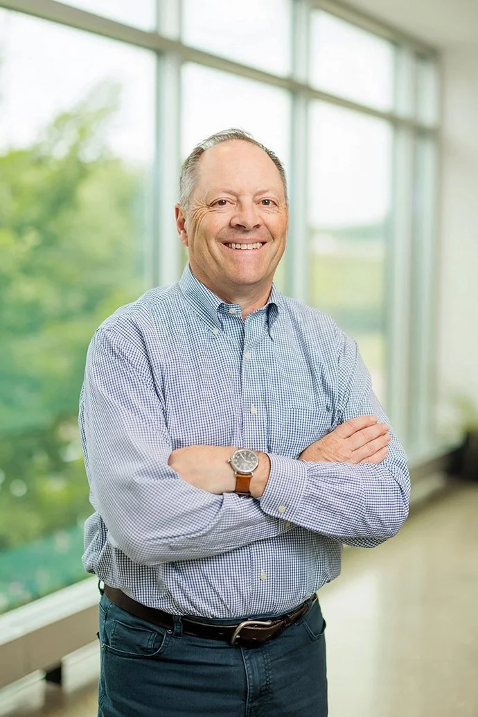 A smiling middle-aged man in a blue checkered shirt with arms crossed, standing in a bright room with large windows and a view of green trees outside.