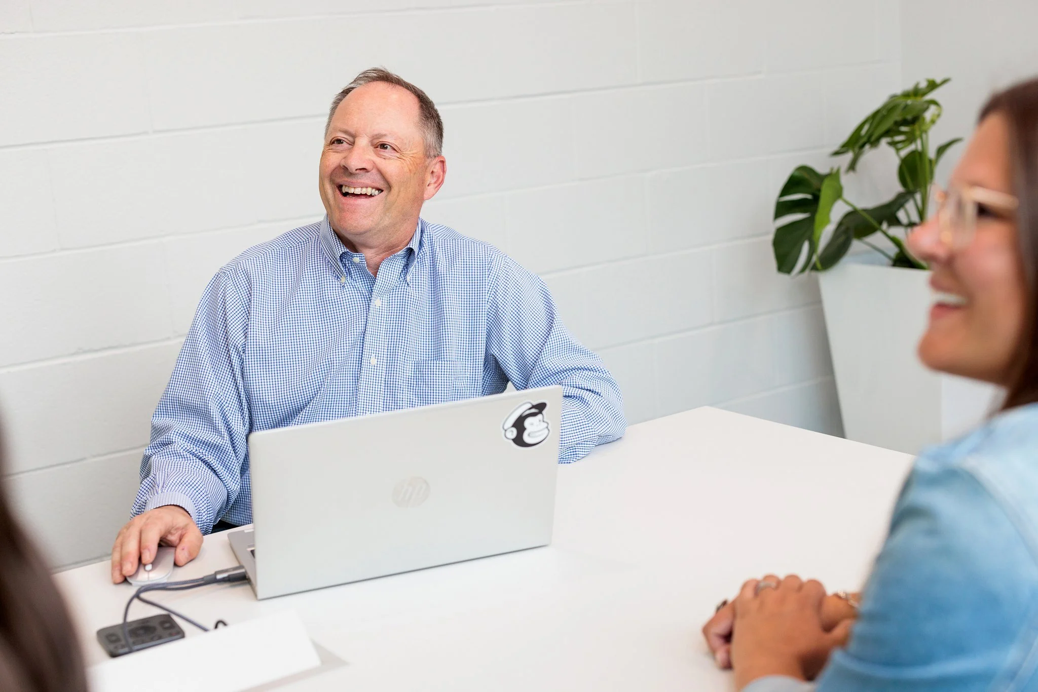 Three people sitting at a white table in a bright, clean room with white brick walls. One man with short gray hair and a checkered shirt is smiling and using a laptop. Two women with glasses, one with brown hair on the right and partially visible, the other with long hair in the foreground, are listening, smiling, and interacting.