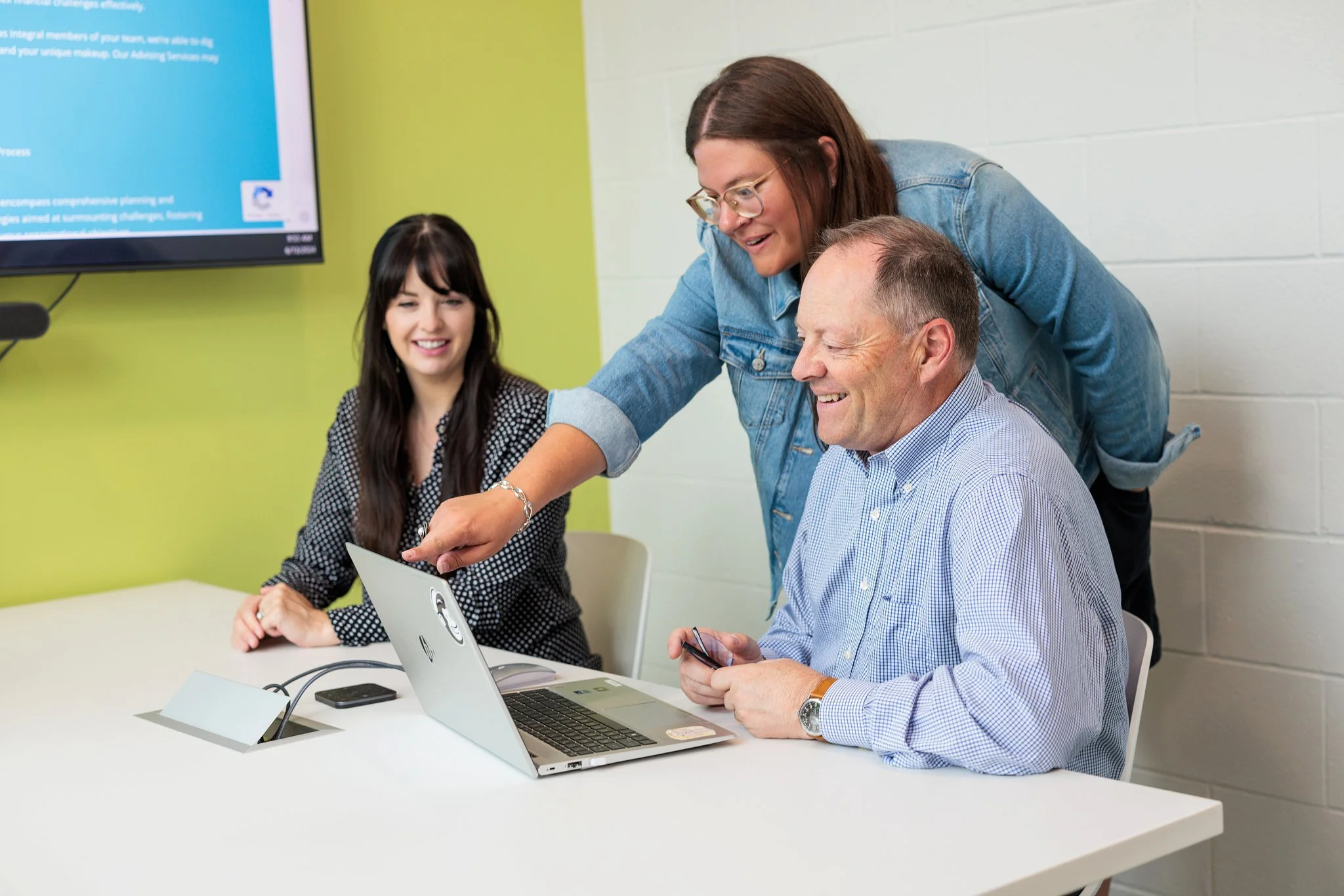 Three people in a meeting room, two women and one man, looking at a laptop and smiling, with a presentation screen in the background.