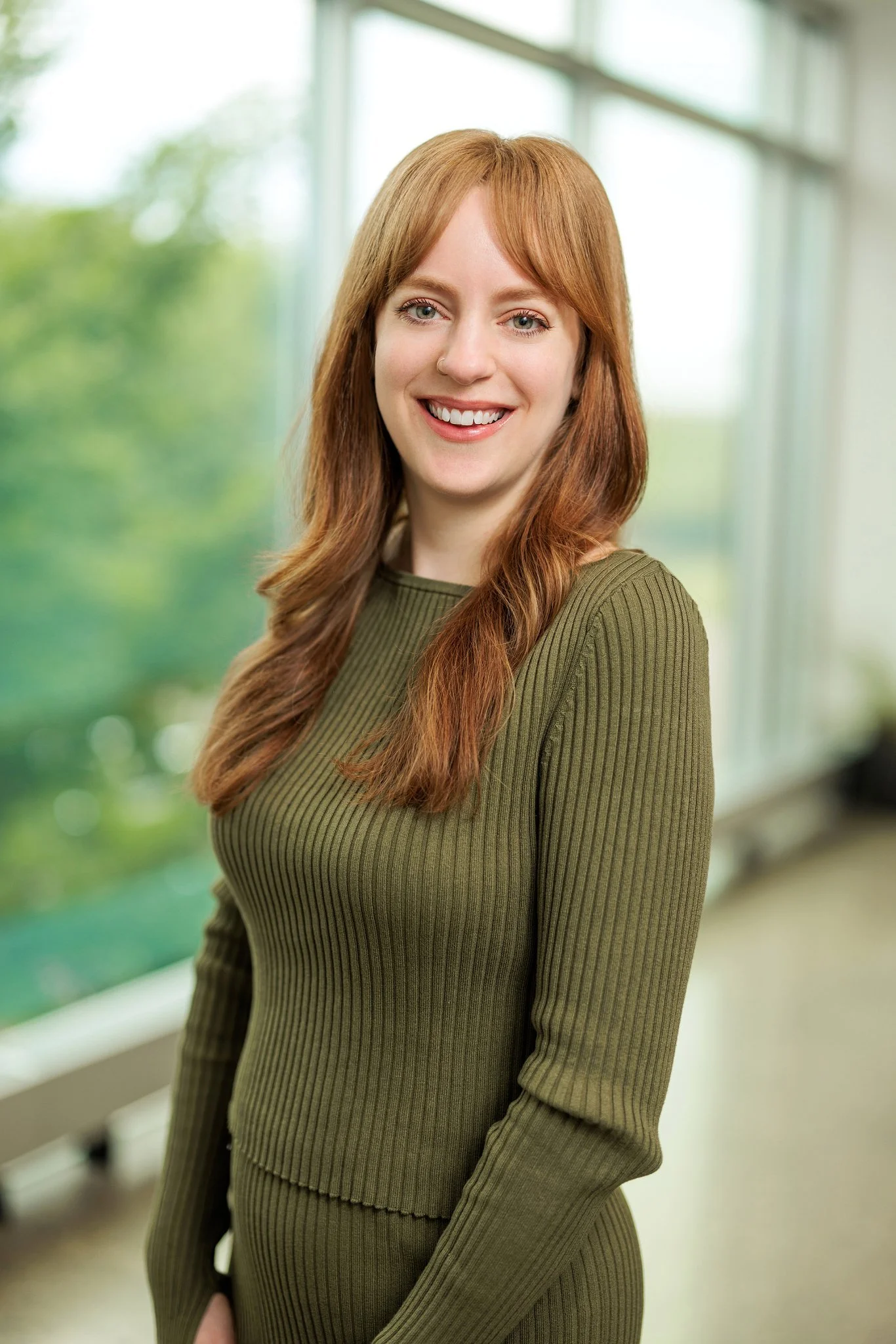 A young woman with red hair and a nose piercing smiling indoors with large windows and green trees outside.