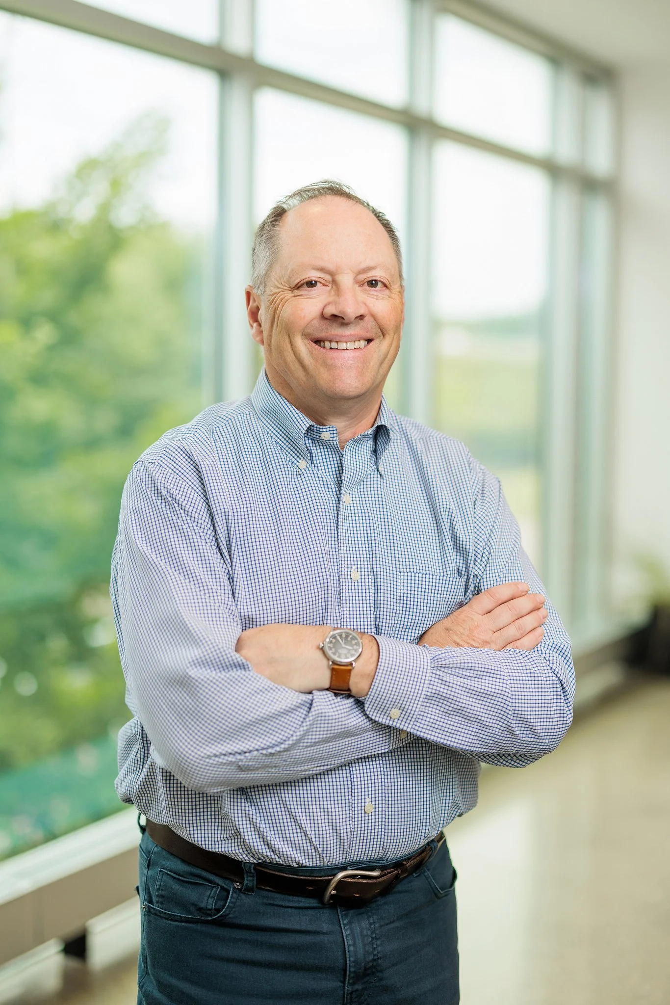 A smiling middle-aged man in a blue checkered shirt with arms crossed, standing in a bright room with large windows and a view of green trees outside.