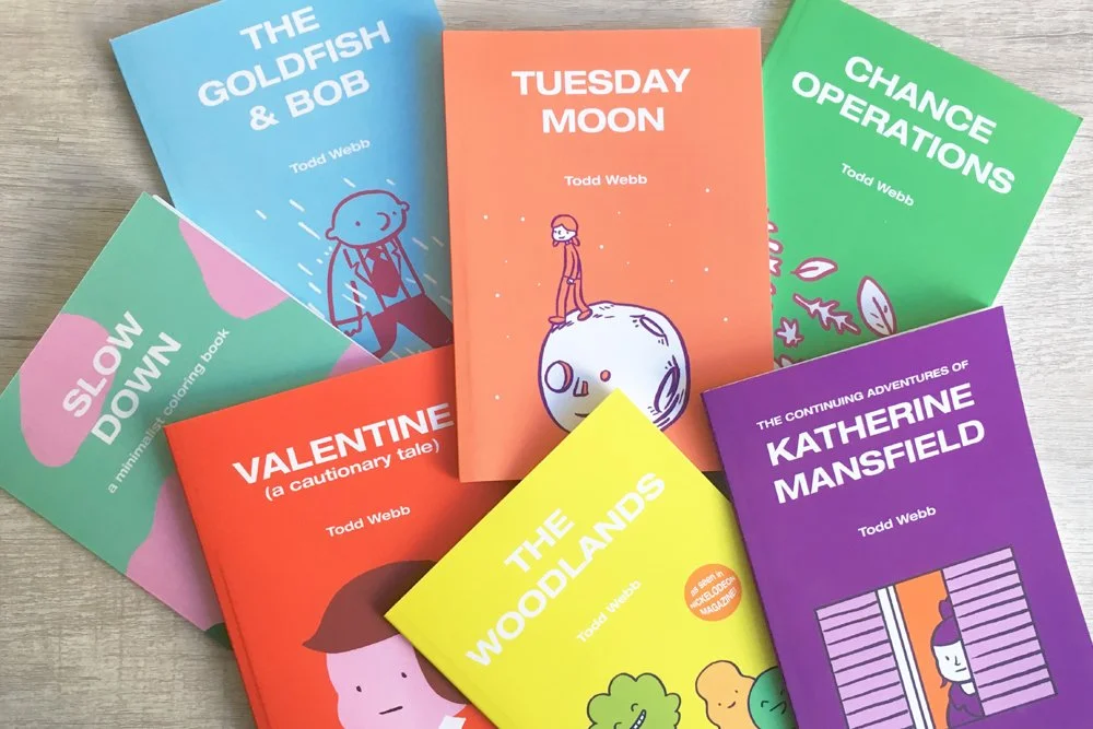 Collection of colorful books by Todd Webb, including titles 'The Goldfish & Bob,' 'Tuesday Moon,' 'Chance Operations,' 'Slow Down,' 'Valentine,' 'The Woodlands,' and 'The Adventures of Katherine Mansfield,' arranged on a wooden surface.