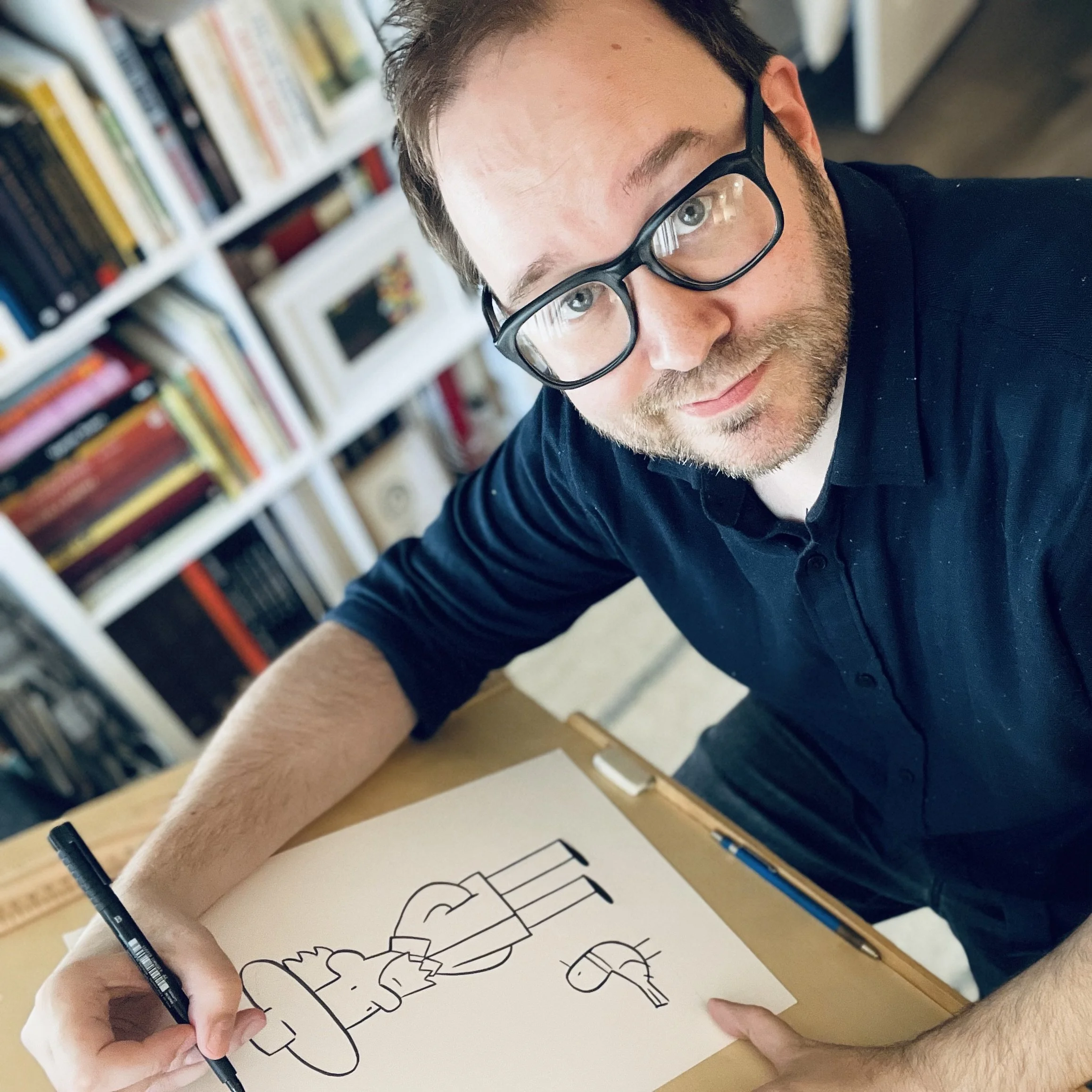 A portrait of cartoonist and musician Todd Webb sitting at his drawing board in a studio filled with books. He is drawing a large picture of his comic strip characters from The Poet.