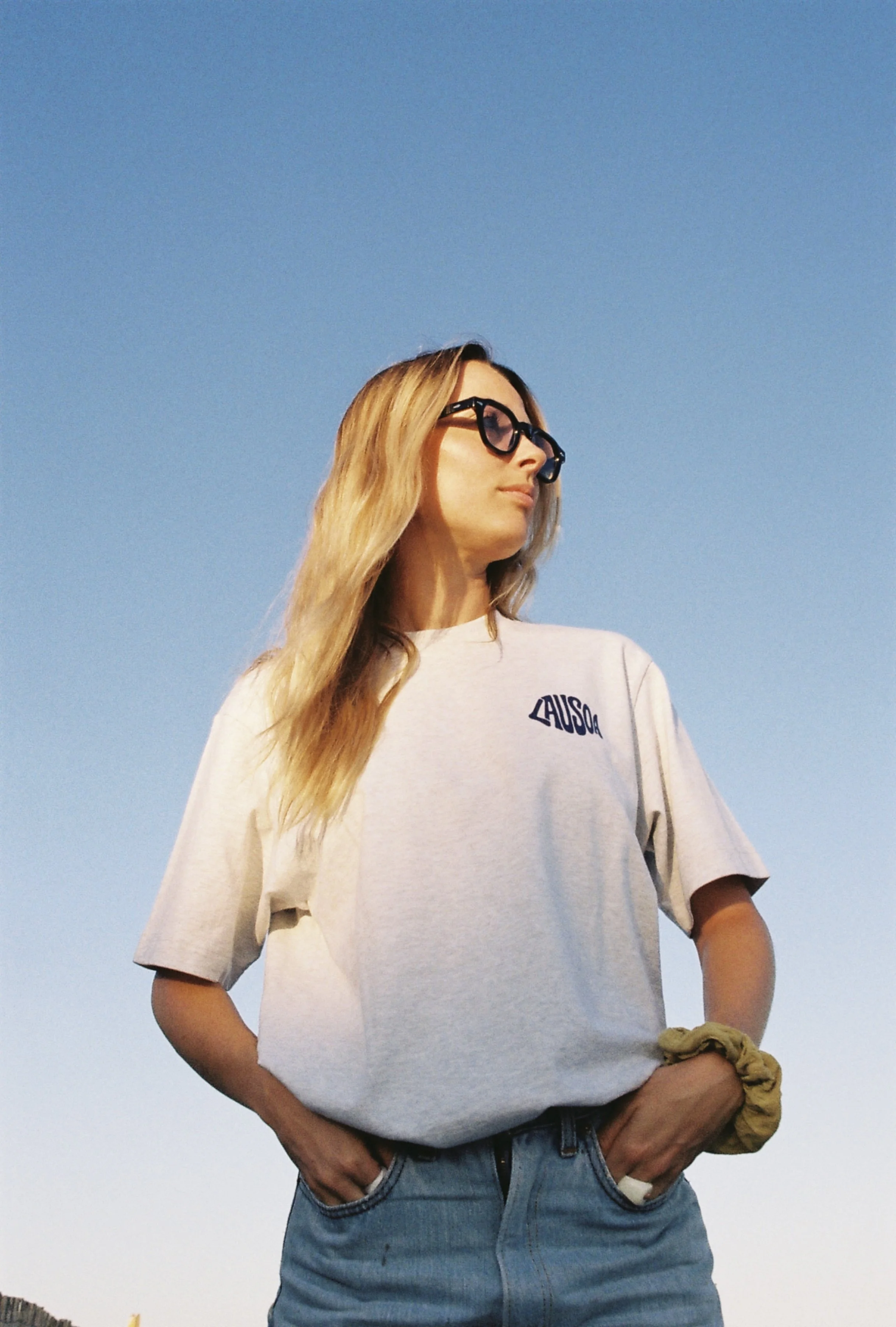 Jeune femme avec lunettes noires, cheveux longs et blonds, portant un t-shirt beige avec une inscription, dans un décor extérieur sous un ciel bleu.