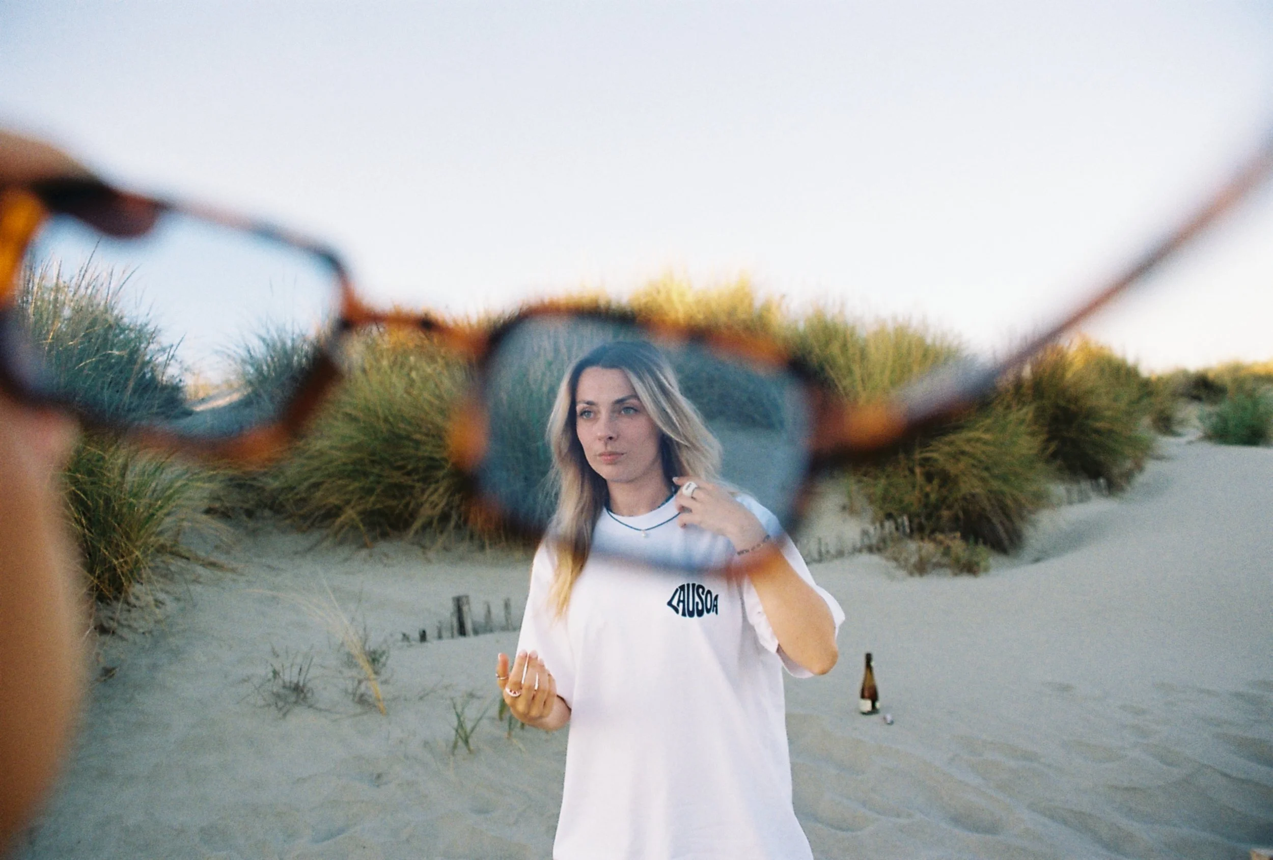Une femme blonde vue dans un miroir de soleil, portant un t-shirt blanc dans un environnement de dunes et de végétation, avec une bouteille et un verre en arrière-plan.