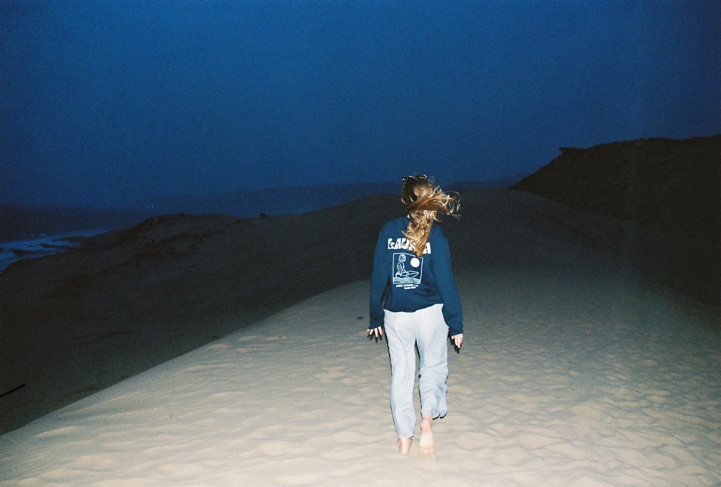 Femme marchant pieds nus sur une dune de sable lors du coucher ou lever du soleil, avec le ciel sombre et des collines en arrière-plan.