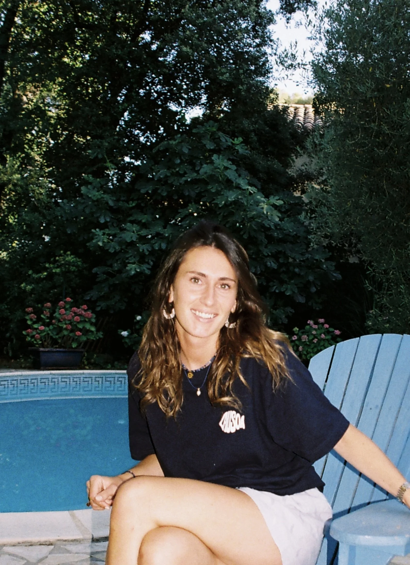 Jeune femme assise sur une chaise bleue à côté d'une piscine, portant un t-shirt noir, des boucles d'oreilles et un collier, avec un jardin luxuriant en arrière-plan.