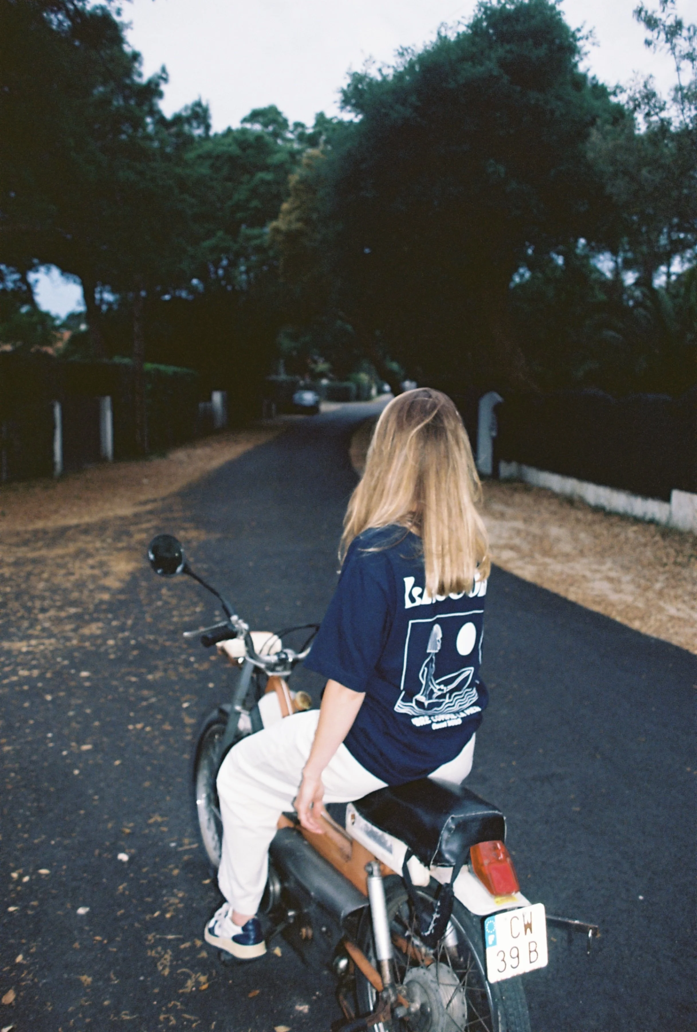 Jeune femme en t-shirt bleu assise sur un vélo électrique sur une route bordée d'arbres, en soirée calme.