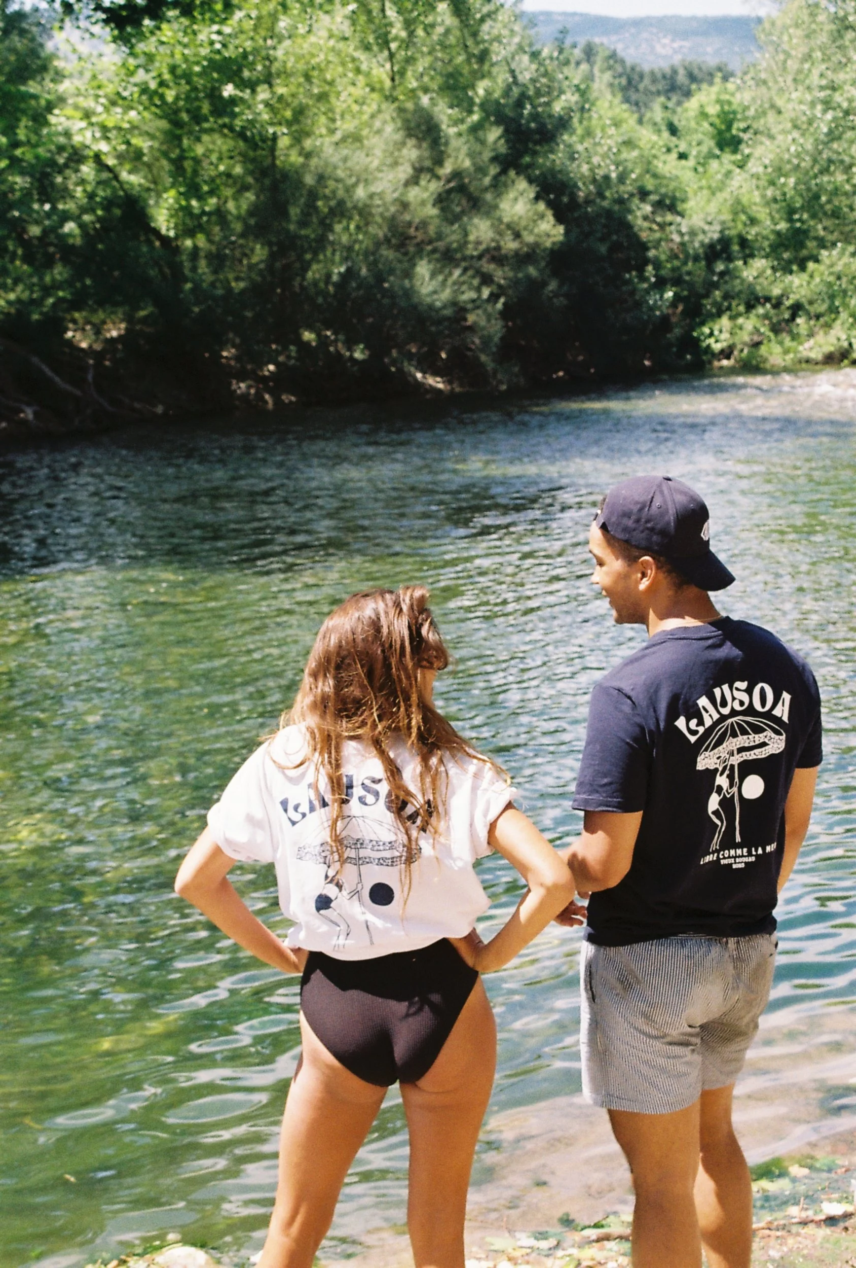 Deux jeunes personnes debout au bord d'une rivière, sous un arbre avec beaucoup de feuillage vert, lors d'une journée ensoleillée portant des t-shirts lausoa d'été avec une femme qui ferme un parasol au coucher de soleil sur la plage.