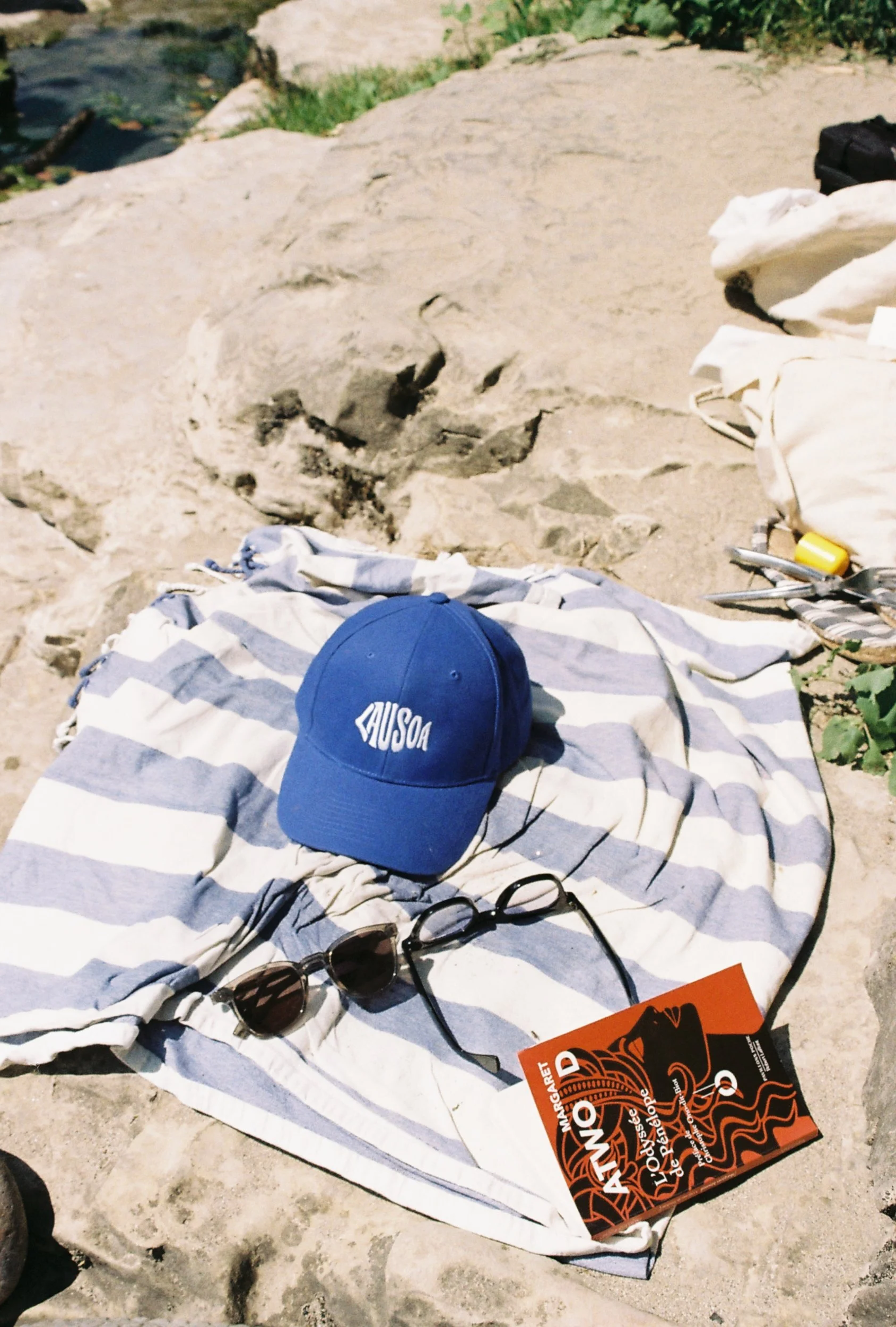 Accessoires de plage sur une serviette rayée, comprenant une casquette bleue avec le mot 'AUSOA', une paire de lunettes de soleil, et un livre de Margaret Atwood. Le décor indique une plage avec sable et rochers.