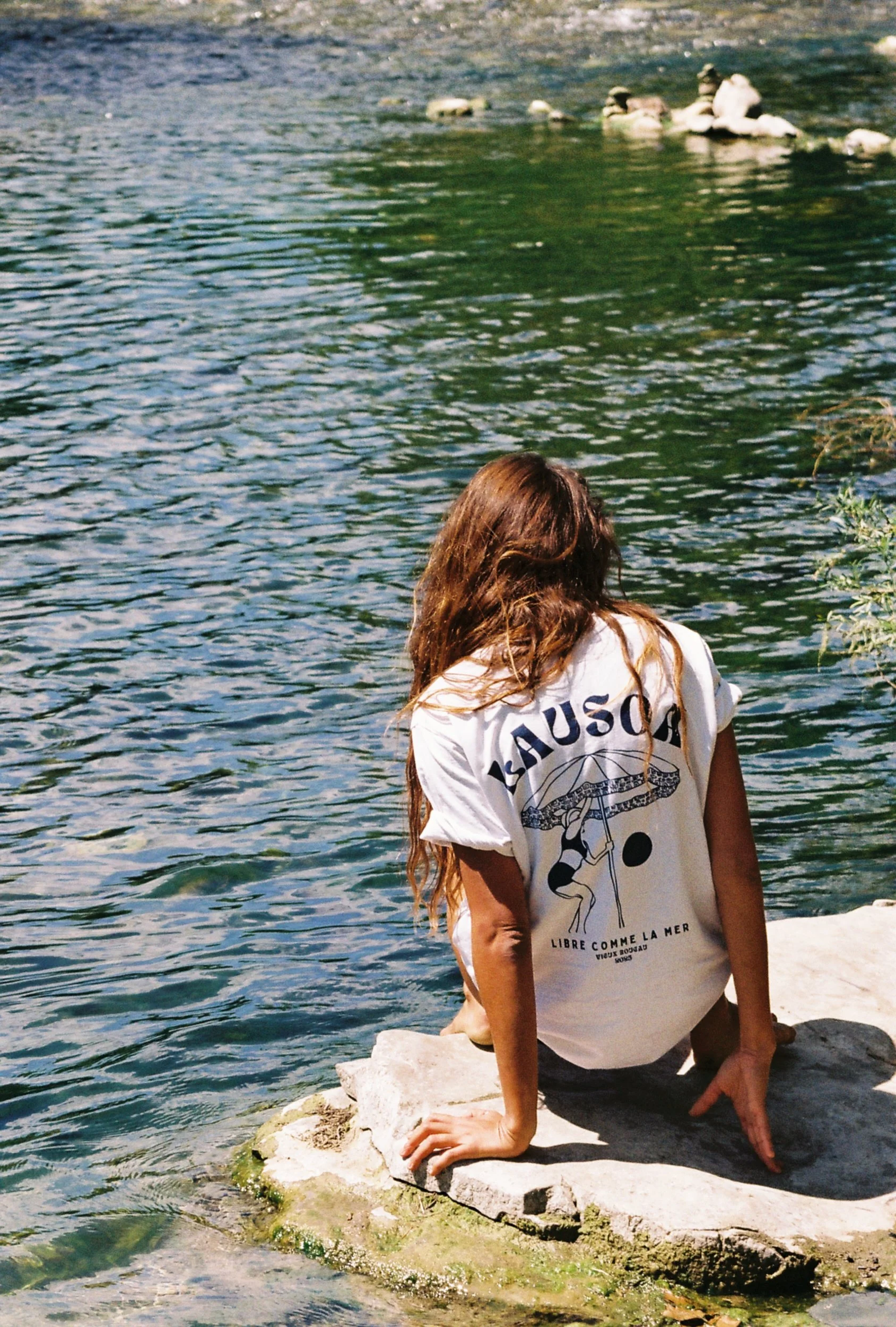 Une personne avec de longs cheveux bruns assise sur une roche au bord de l'eau, portant un t-shirt blanc avec un dessin et le texte 'LAUSOA'.