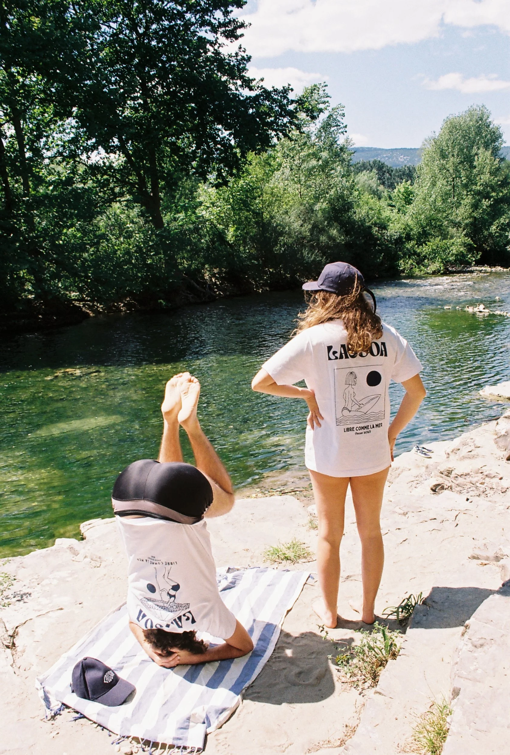 Deux personnes, une femme et un homme, portent des t-shirts blancs avec des motifs et textes en français, près d'une rivière entourée d'arbres verts. La femme se tient debout, regardant la rivière, tandis que l'homme, allongé sur un tapis, fait une p