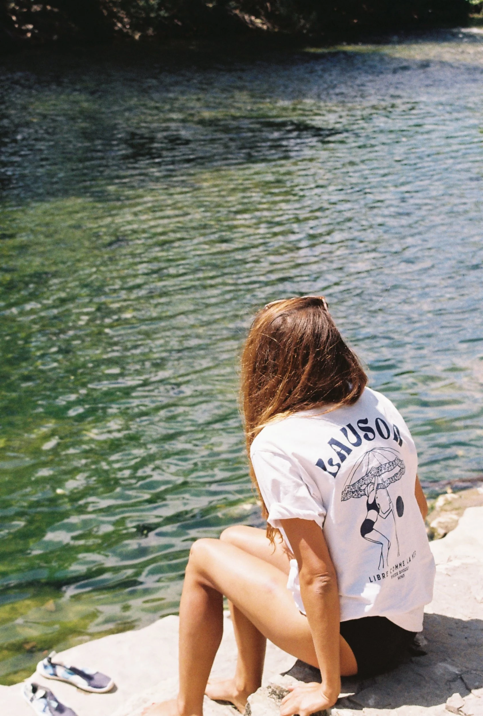 Femme assise sur un rocher au bord de l'eau, portant un t-shirt blanc Lausoa et un short, avec ses cheveux longs bruns cachant son visage, près d'une rivière ou d'un lac.