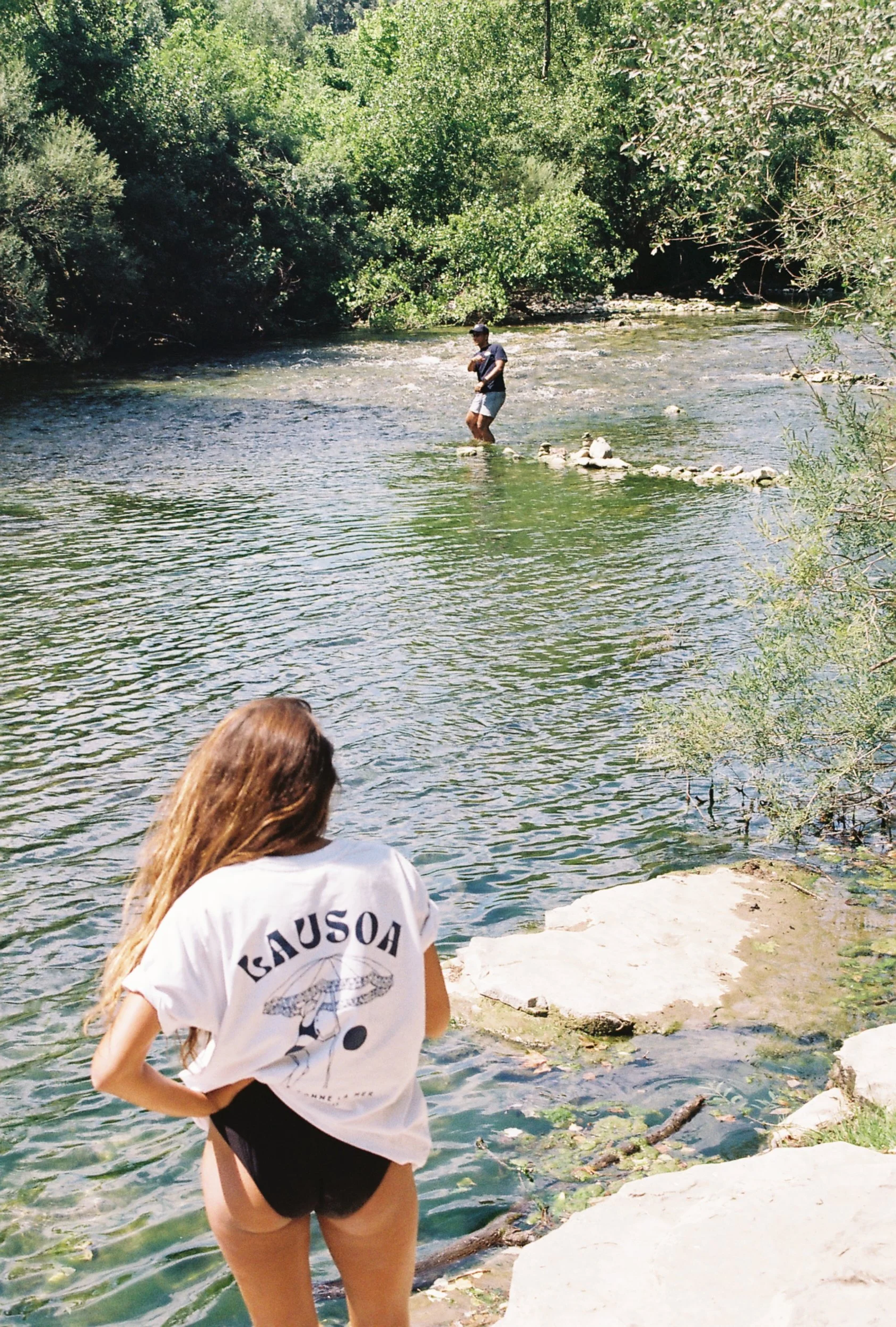 Une femme avec des cheveux longs et bruns, portant un t-shirt blanc avec l'inscription "KAUSOA", regarde la rivière. Au fond, un homme debout dans l'eau proche du rivage porte un t-shirt gris, un short et une casquette, et se tient sur une pierre dan