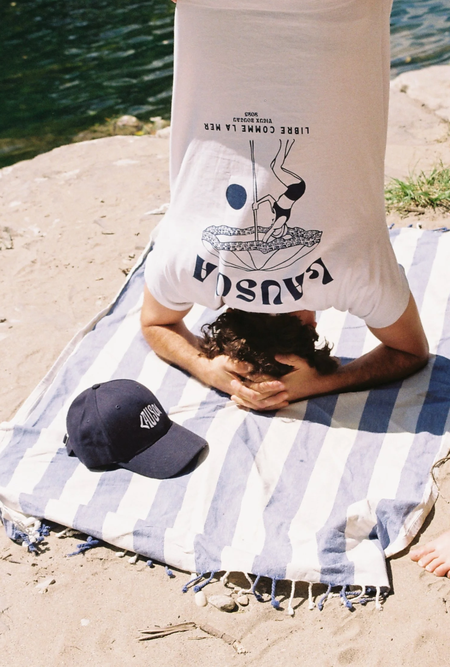 Une personne pratique le yoga ou la méditation sur une serviette rayée en face de l'eau, avec une casquette LAUSOA noire posée sur la serviette et un t-shirt lausoa avec comme motif une femme qu ferme son parasol au coucher de soleil 