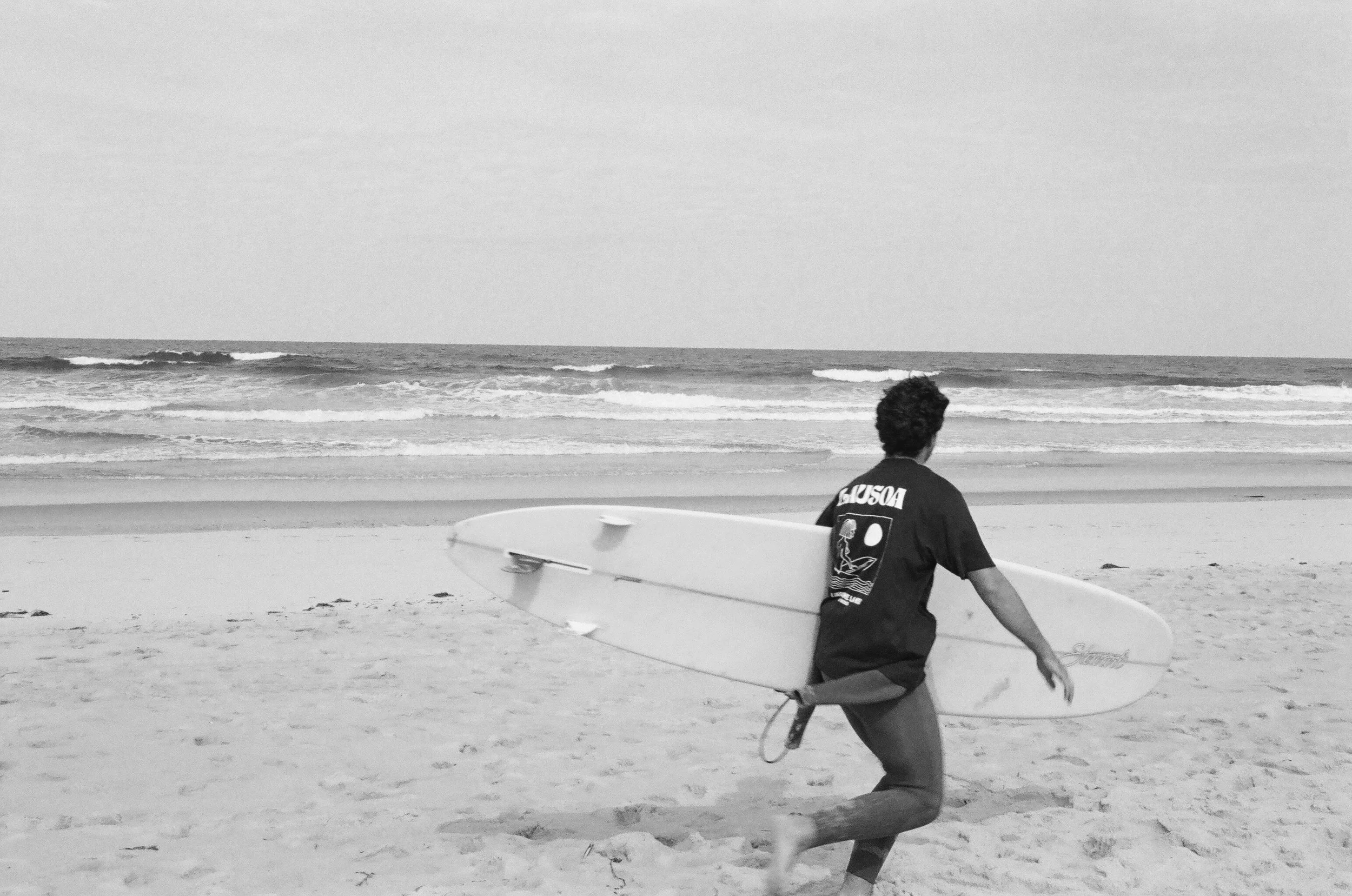 Jeune homme marchant sur la plage avec une planche de surf, vue de derrière, mer et ciel nuageux en arrière-plan.