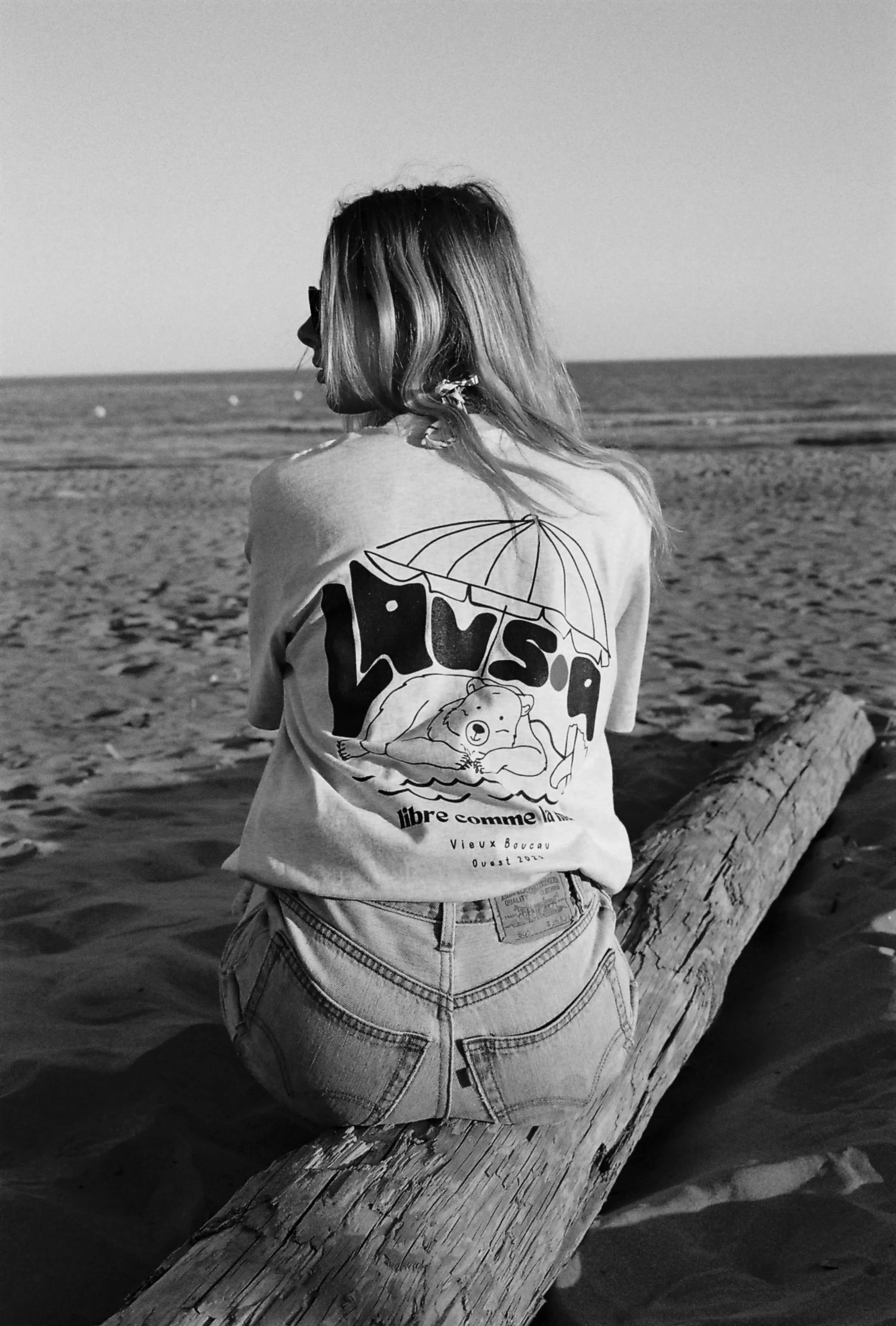 Jeune femme blonde assise sur un bois flotté, regardant vers la mer, portant un t-shirt gris avec un dessin d'ours et un parasol, à la plage au coucher du soleil.