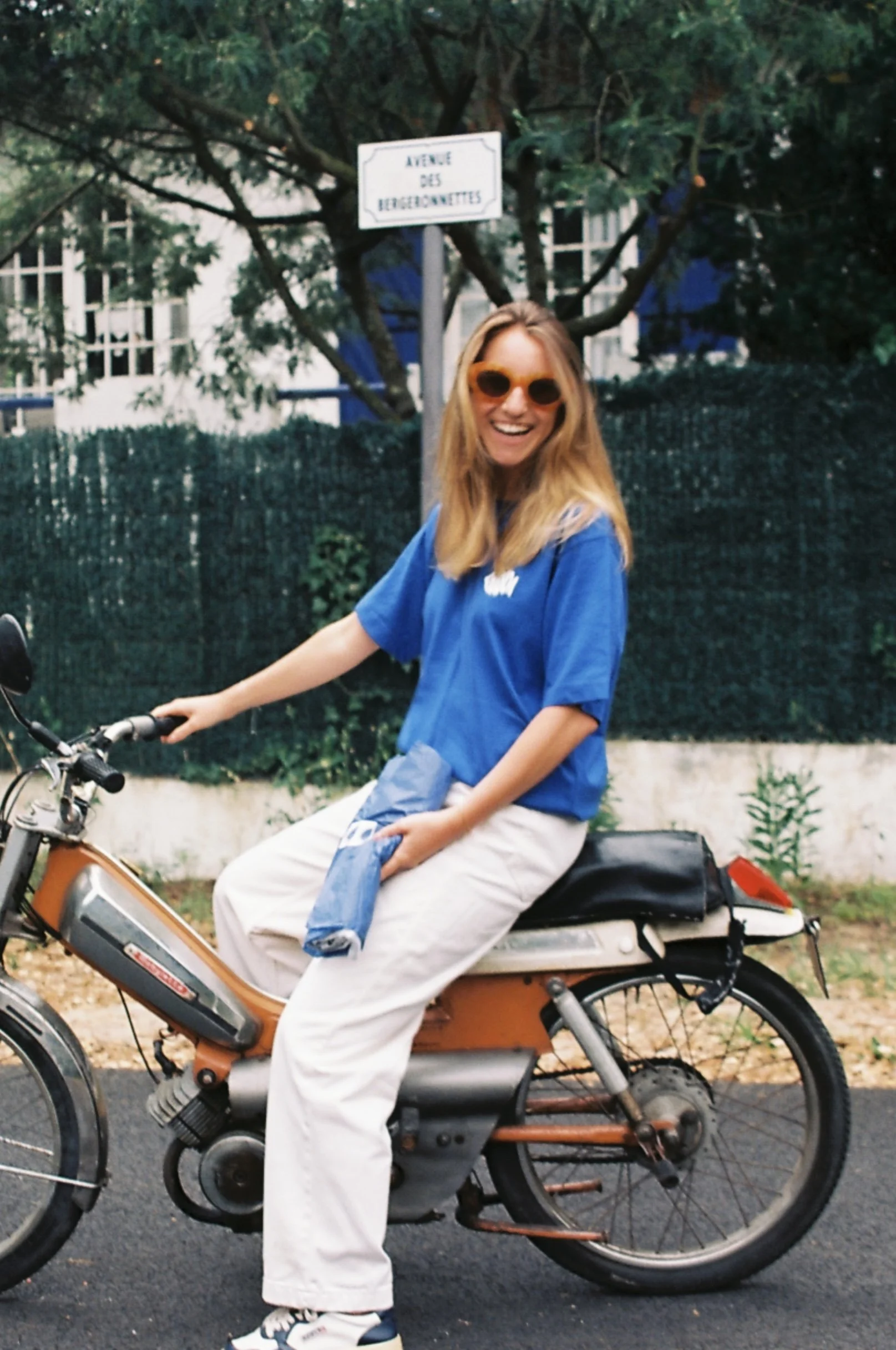 Jeune femme souriante avec des lunettes orange assise sur une moto orange, en arrière-plan un arbre et une enseigne de rue "Avenue des Berquinette".