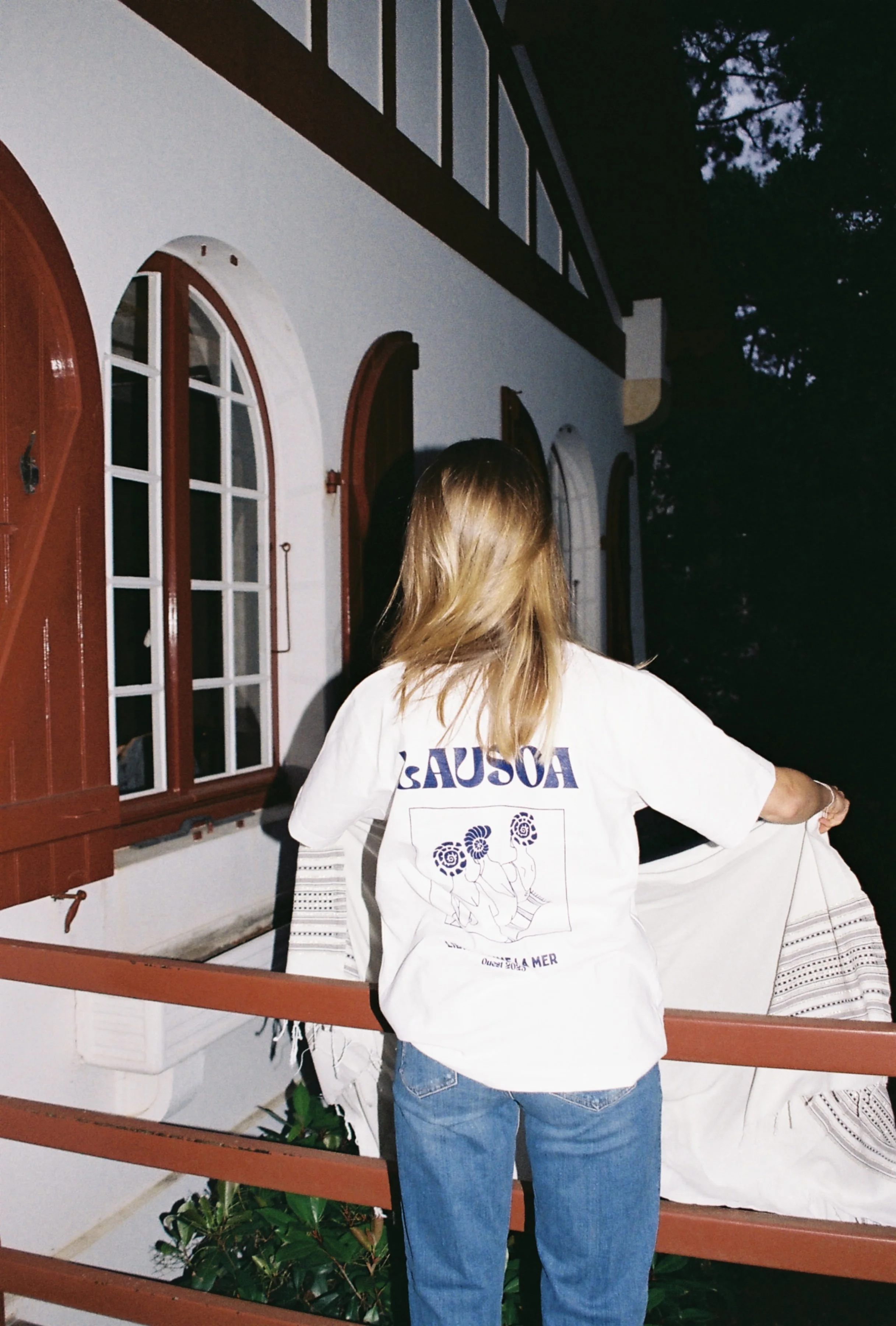 Femme de dos tenant un rideau blanc devant une maison blanche avec fenêtres à volets rouges, la nuit