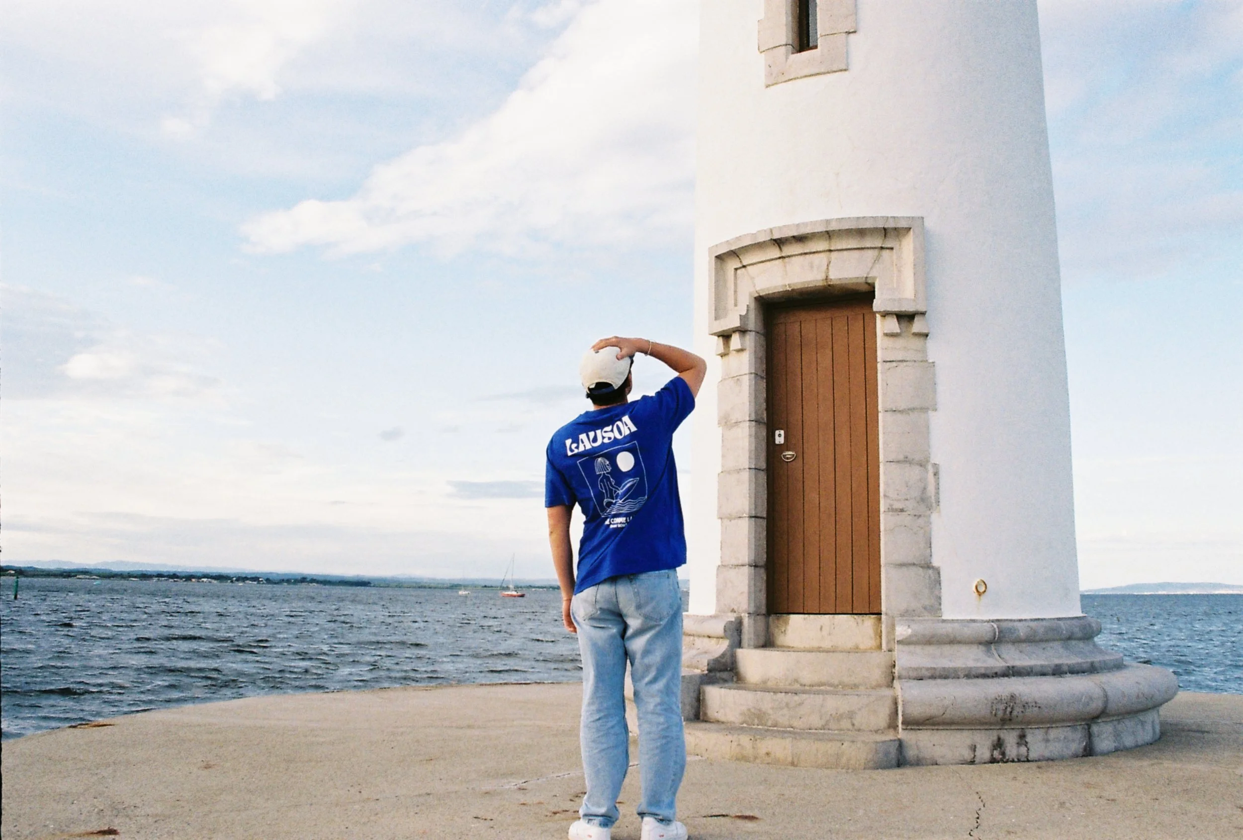 Personne vue de dos portant un t-shirt bleu avec textes et dessin, sans nom, en regardant un phare blanc près de la mer.