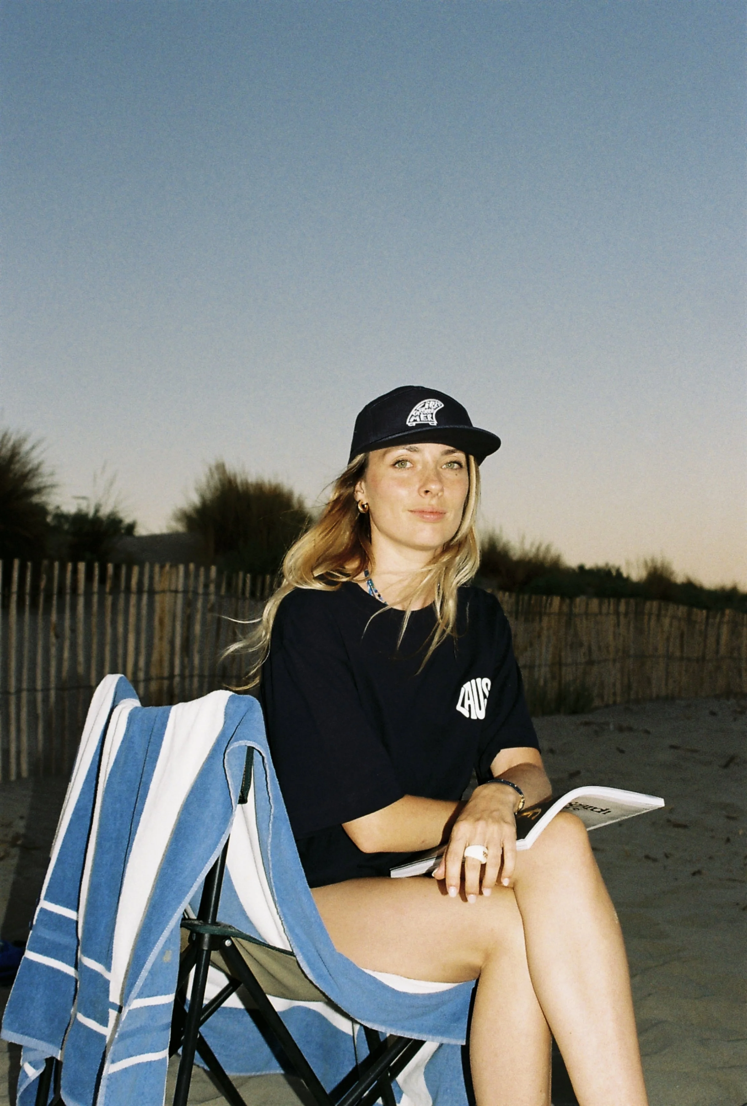Une femme assise sur une chaise pliante à la plage lors du coucher du soleil, portant une casquette noire, un t-shirt noir, et tenant un livre ou magazine.