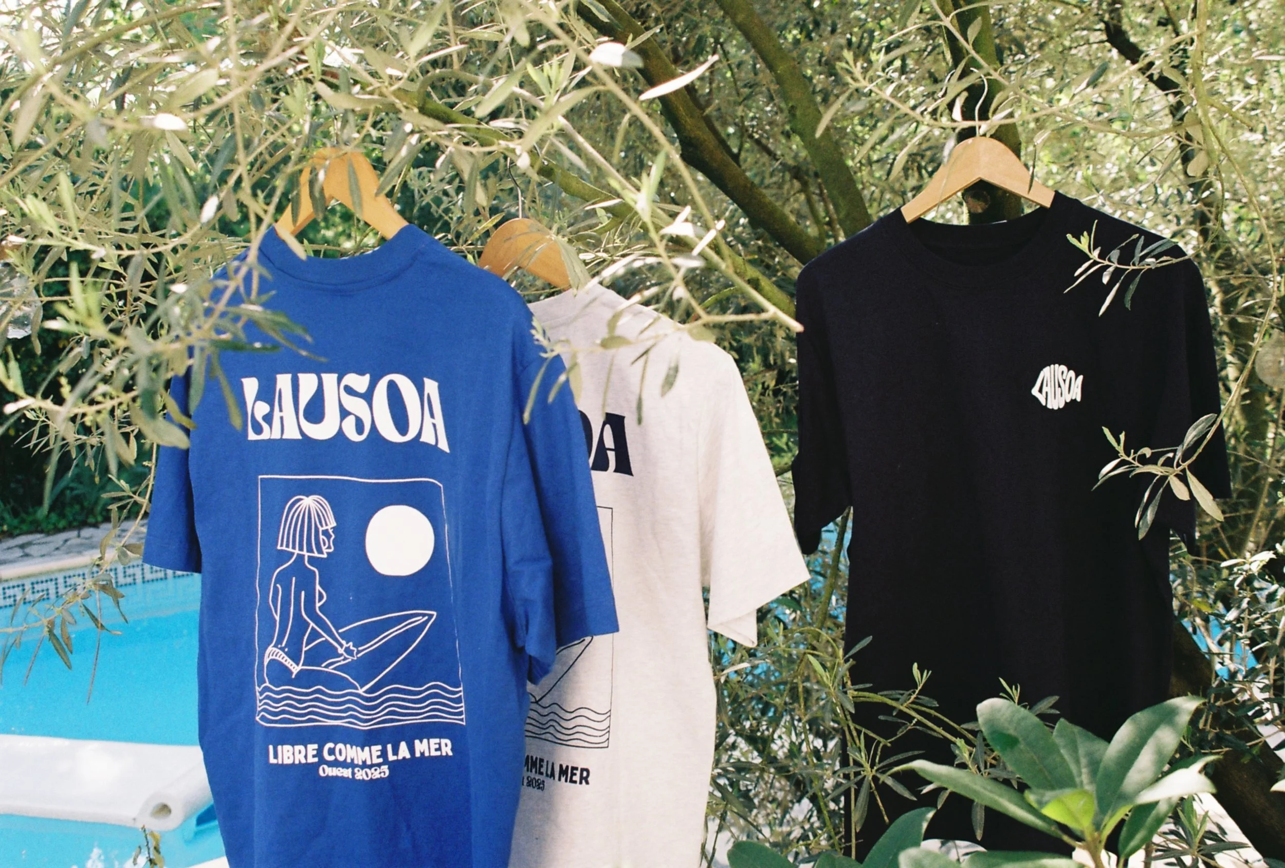 Trois t-shirts suspendus sur des cintres dans un environnement naturel avec des arbres, un fond de piscine, et des branches d'olivier. Le t-shirt bleu à gauche affiche une illustration d'une femme assise sur une planche de surf avec un soleil et la p