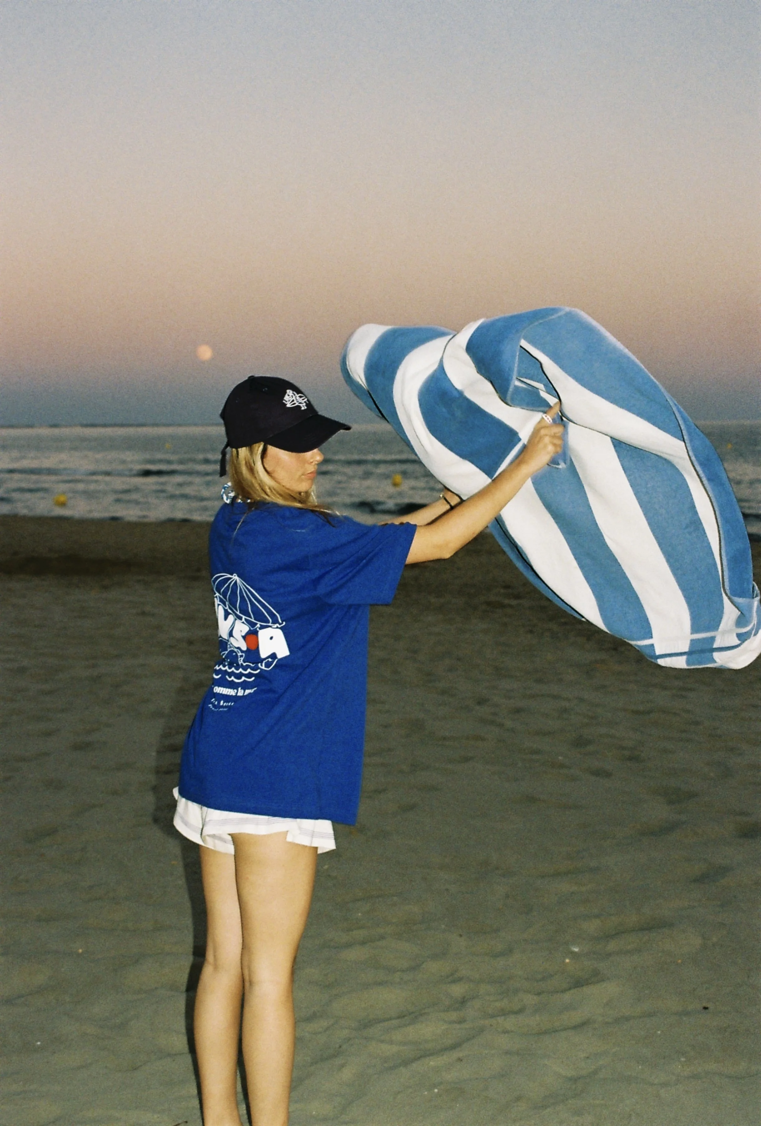 Jeune femme sur la plage, déployant un parasol rayé bleu et blanc, au coucher du soleil.