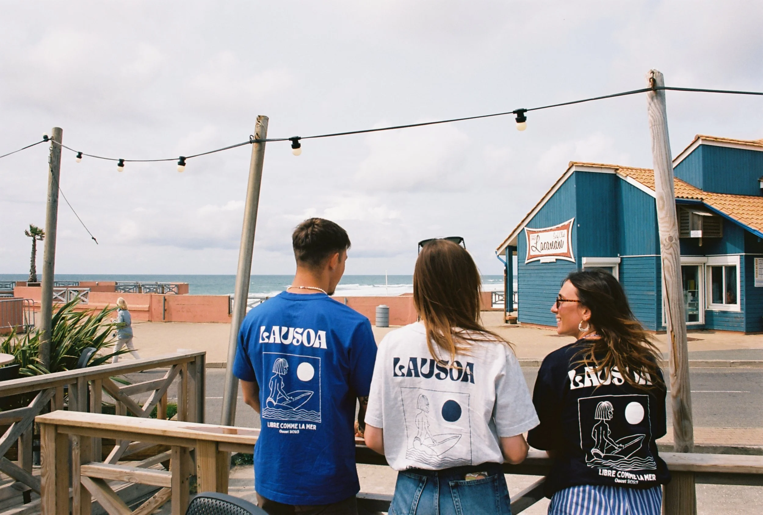 Trois jeunes portant des t-shirts avec le mot 'LAUSOA' et une illustration de femme sur la plage, avec le sable, la mer, une maison bleue et un ciel nuageux en arrière-plan.