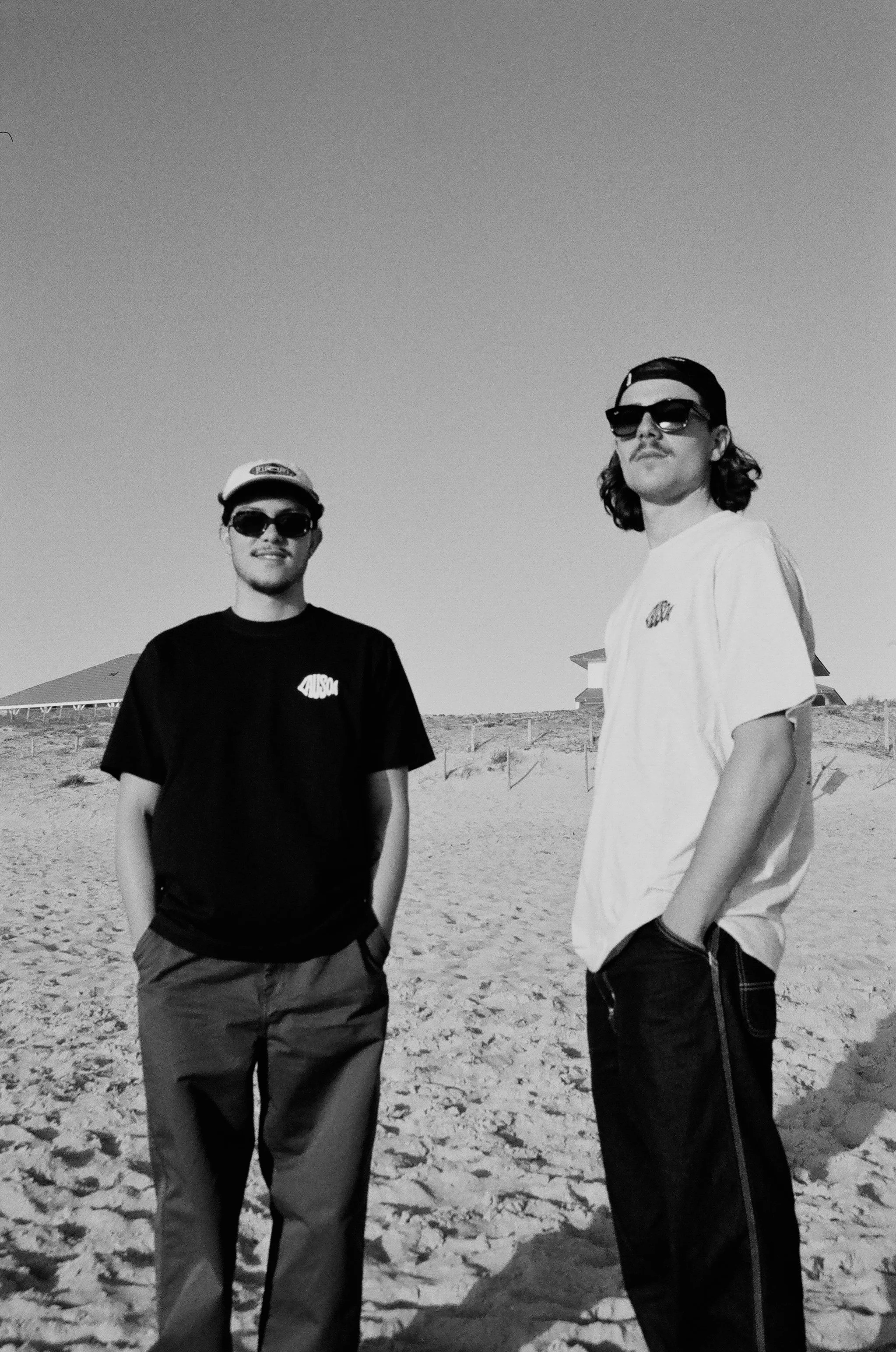 Deux jeunes hommes portant des lunettes de soleil, debout sur une plage, en noir et blanc.