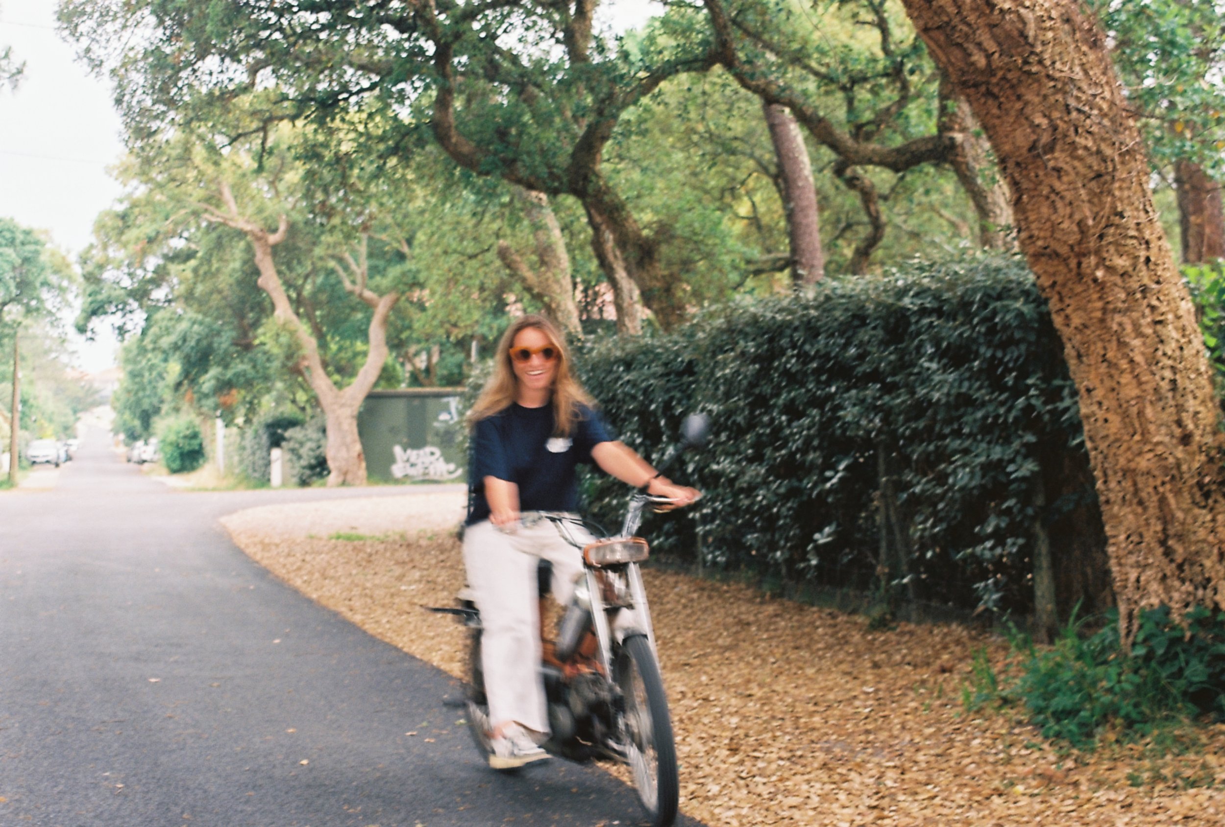 Une femme souriante portant des lunettes de soleil, chevauchant une bicyclette sur une voie pavée bordée d'arbres verts, dans une ambiance en plein air et décontractée.