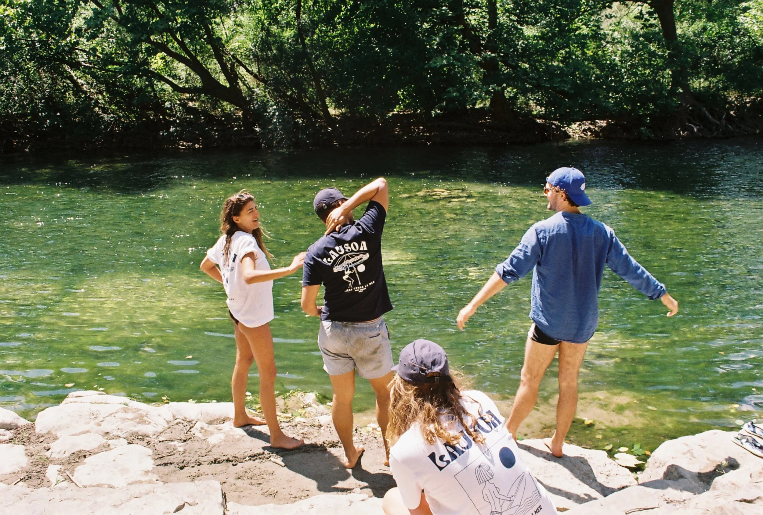 Groupe de jeunes au bord d'une rivière, en vacances, riant et discutant dans un cadre naturel avec des arbres verdoyants en arrière-plan.