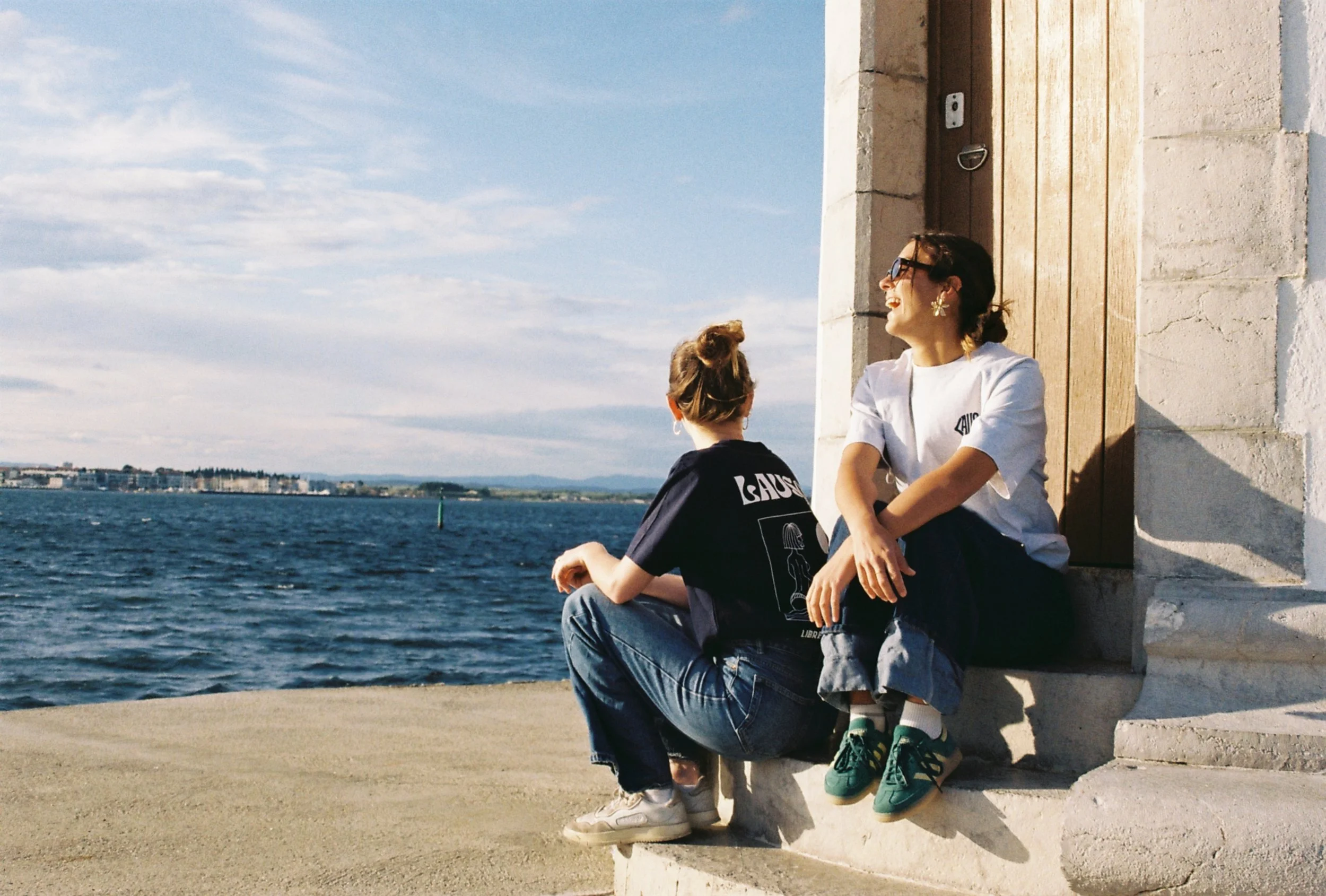 Deux femmes assises près de l'eau, souriant et discutant, avec un bâtiment en pierre et une porte en bois derrière elles, dans un cadre ensoleillé