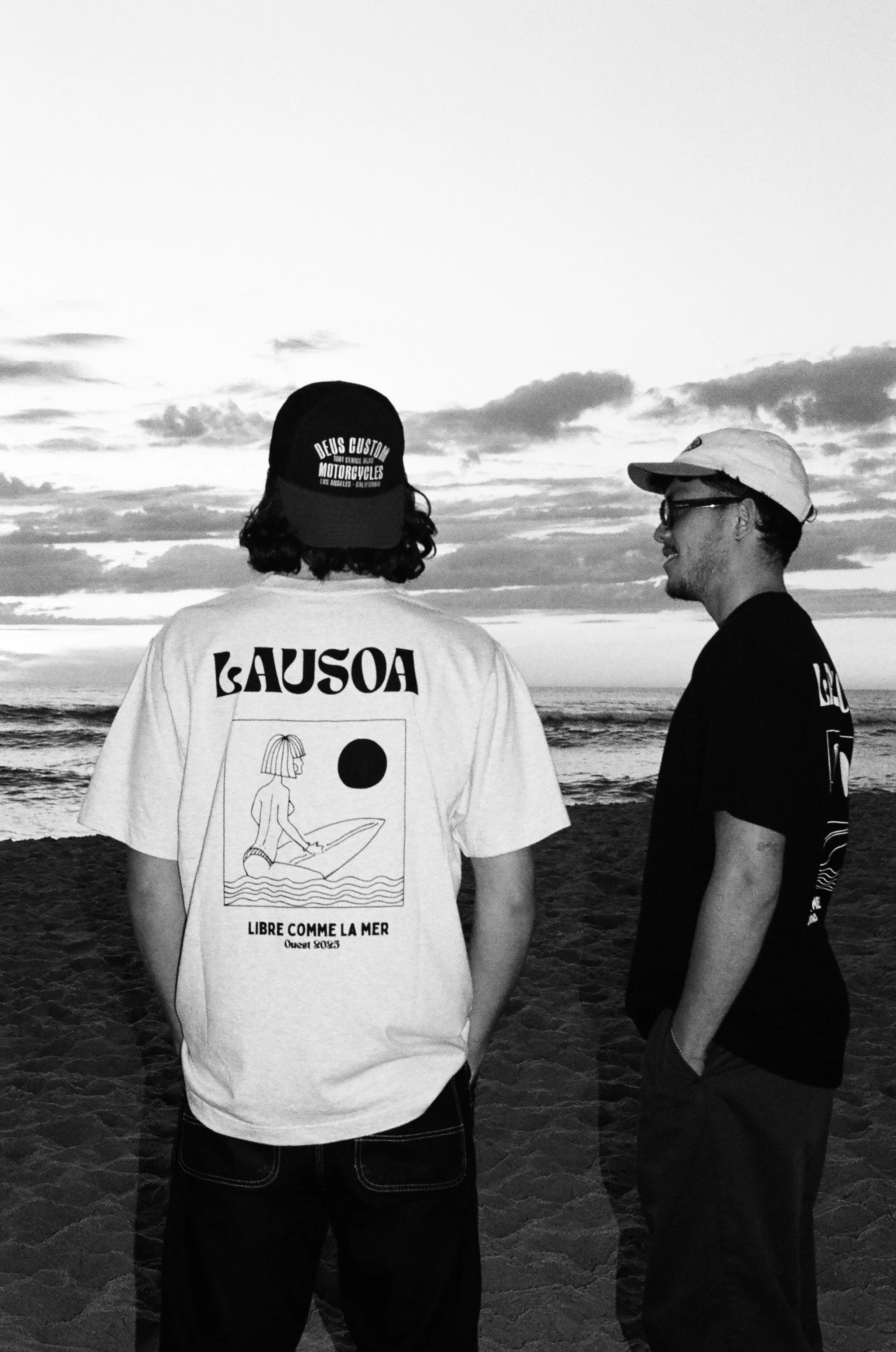 Deux jeunes hommes discutent sur la plage au coucher du soleil, en noir et blanc, portant des casquettes et des t-shirts à motifs.