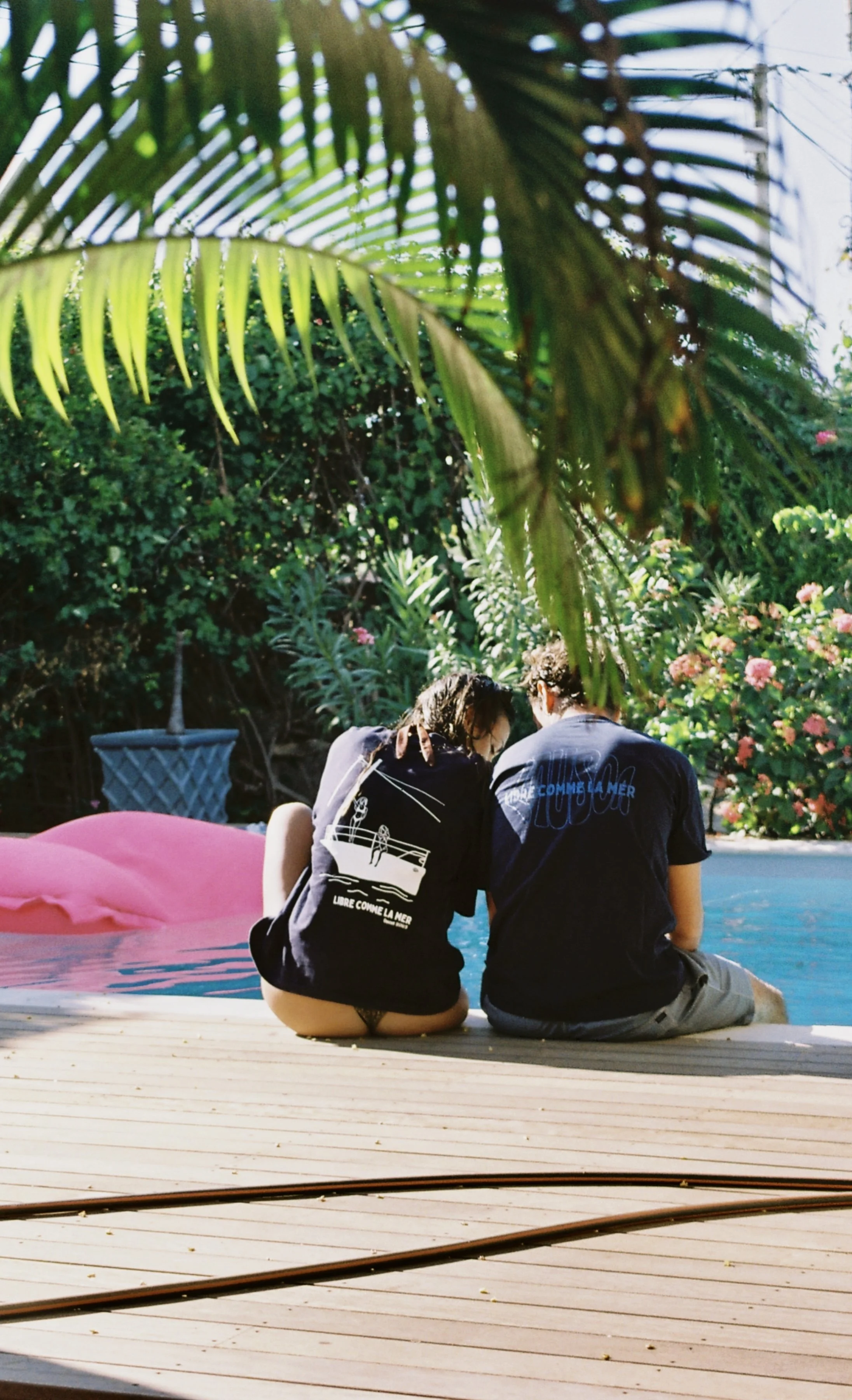 Deux jeunes assis au bord d'une piscine, sous une végétation luxuriante, partageant un moment intime, avec des fleurs roses en arrière-plan.
