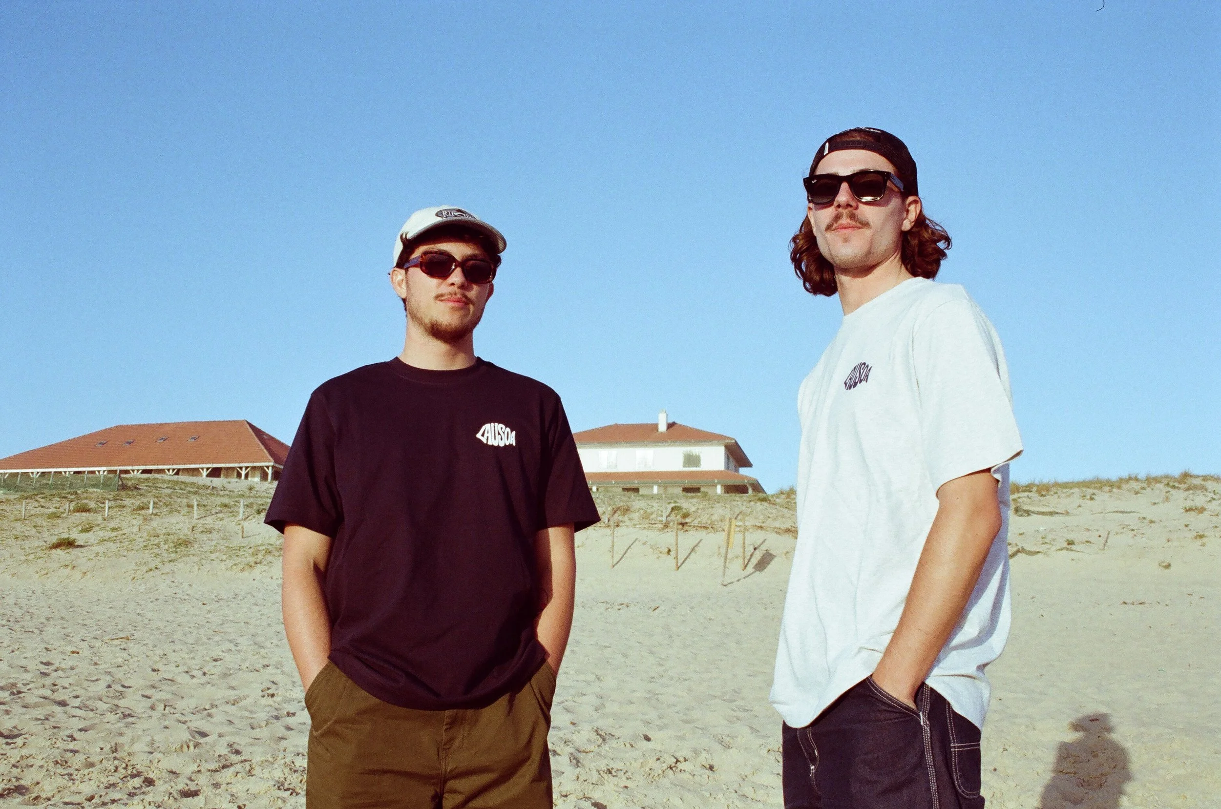 Deux jeunes hommes portant des T-shirts et des lunettes de soleil se tiennent sur une plage avec un bâtiment en arrière-plan, sous un ciel clair.