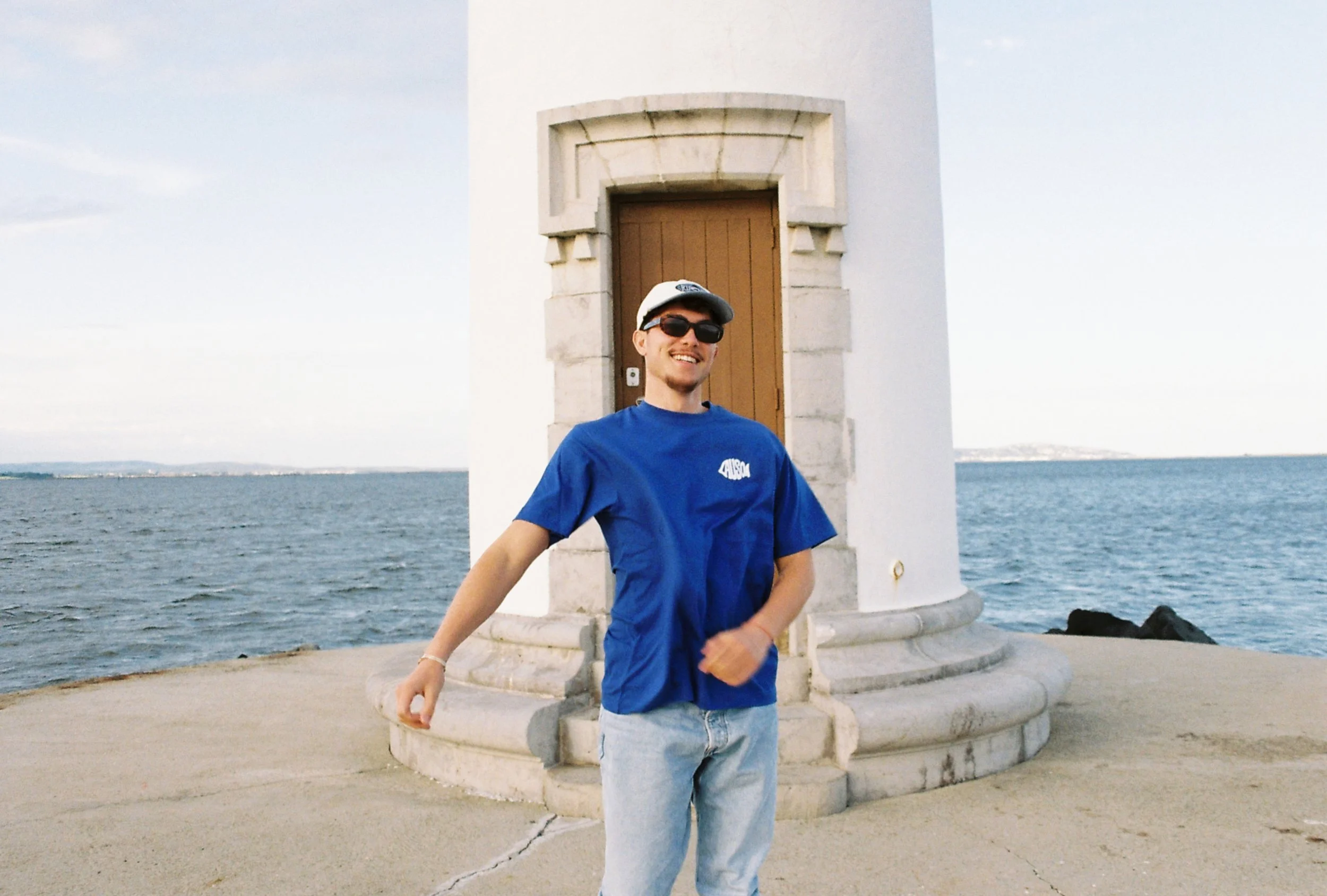 Un homme souriant portant une chemise bleue, des lunettes de soleil et une casquette, debout en face d'un phare près de la mer