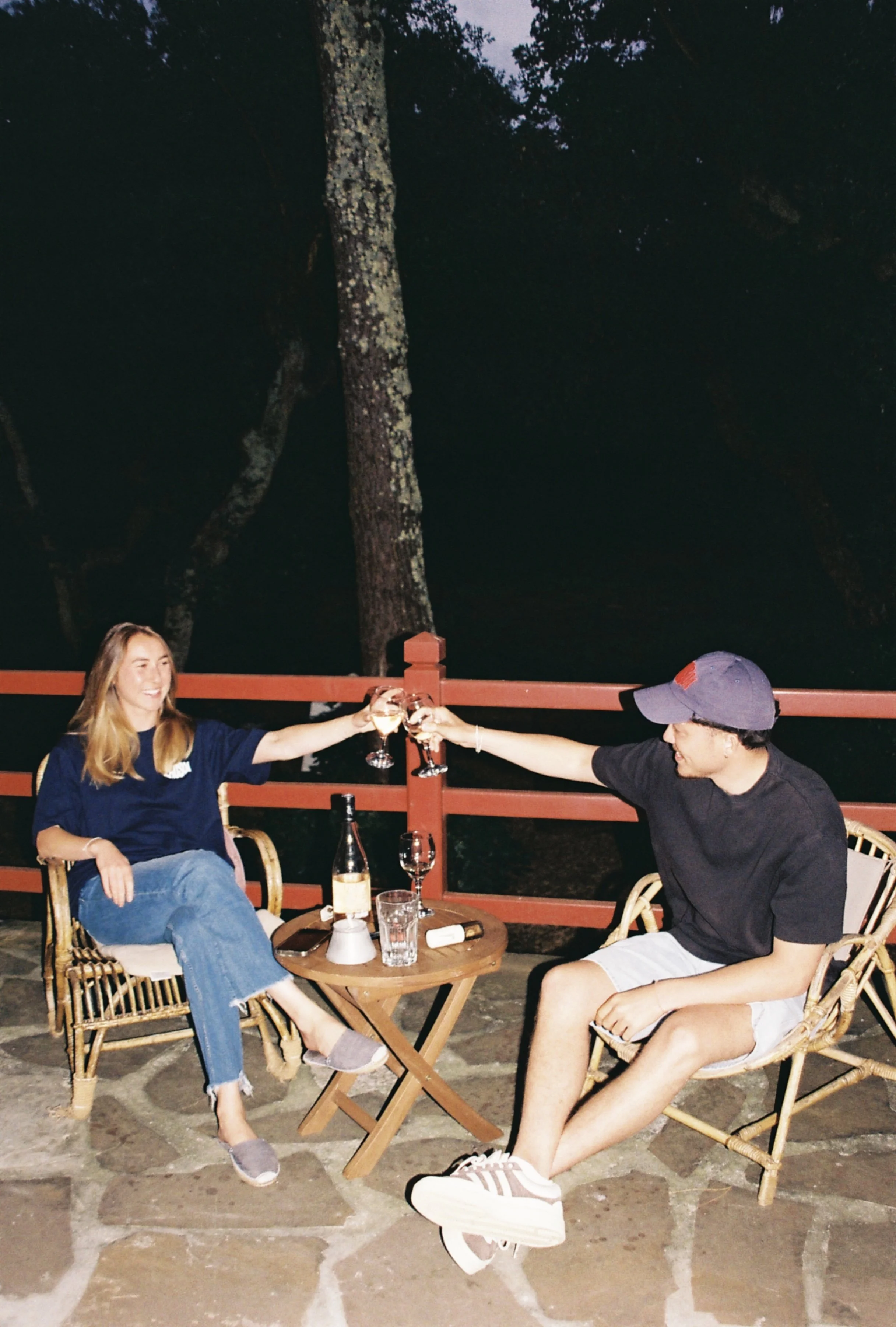 Deux jeunes assis en extérieur, levant leurs verres de vin pour un toast, sur une terrasse en soirée, avec un arbre en arrière-plan.