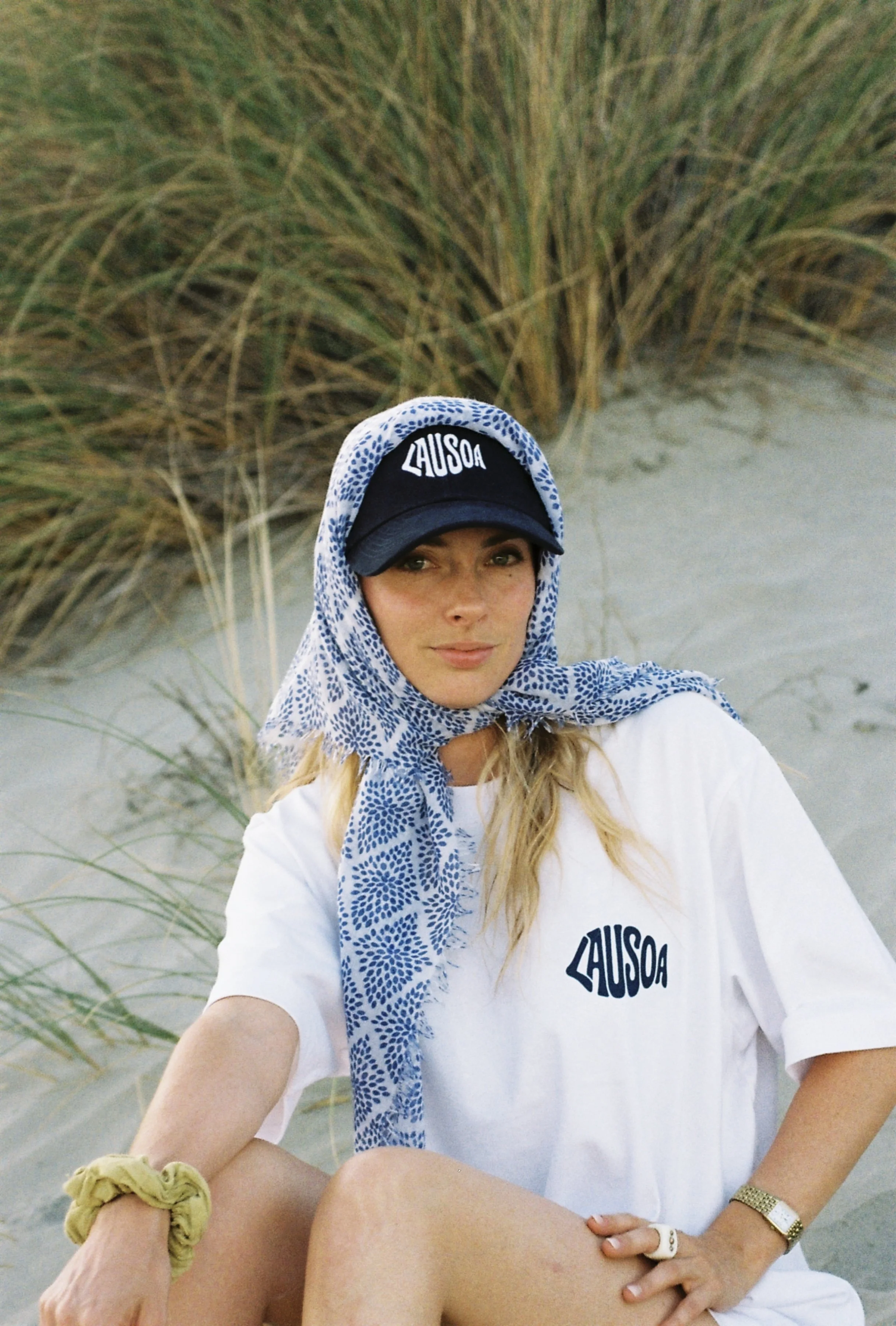 Jeune femme portant un bonnet, une écharpe et un t-shirt avec le mot 'AUSOA' écrit dessus, assise sur la plage avec des plantes dunaires en arrière-plan.