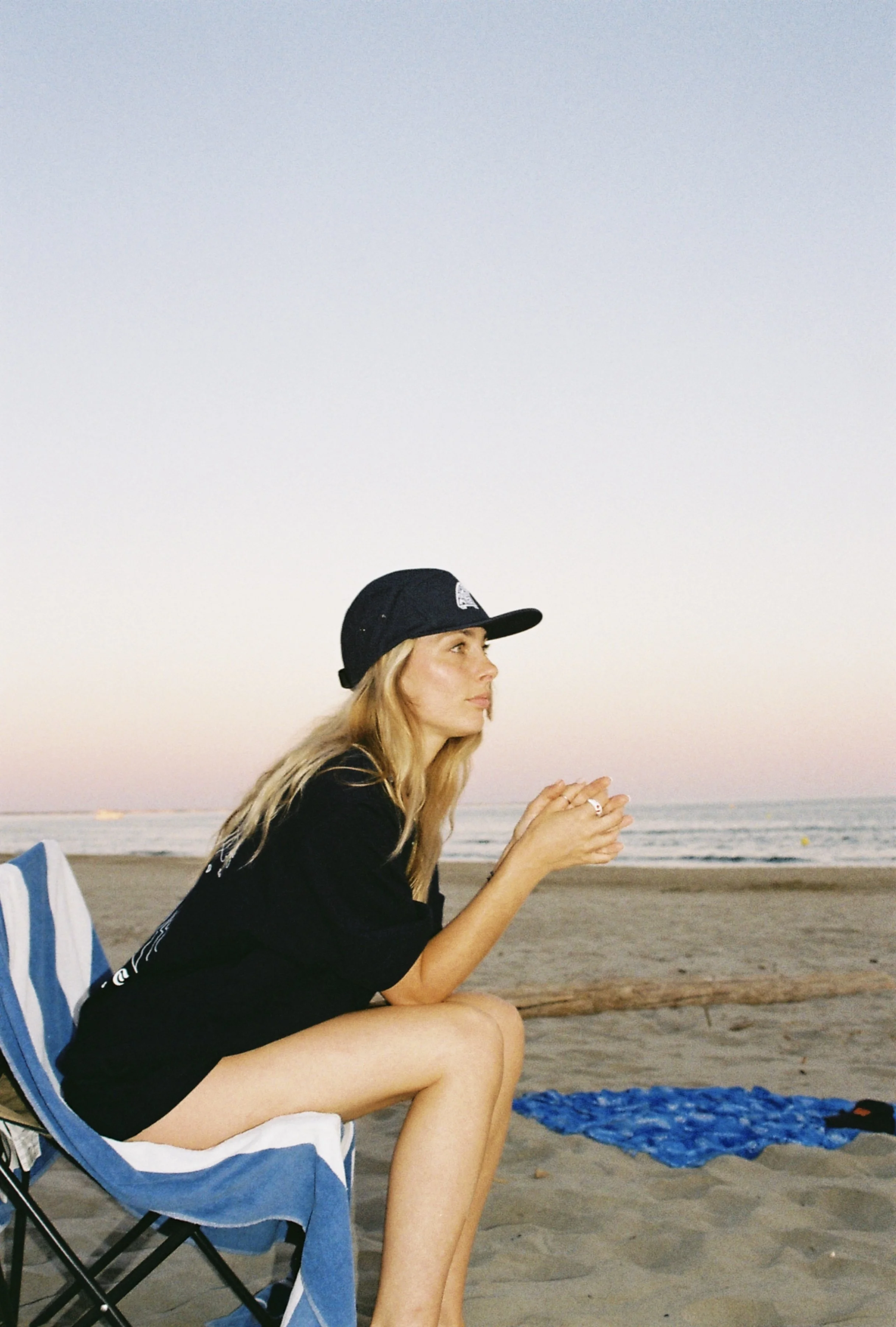 Jeune femme assise sur une chaise de plage en regardant la mer au coucher du soleil, portant une casquette noire et une veste noire, avec un drap de plage à ses côtés.