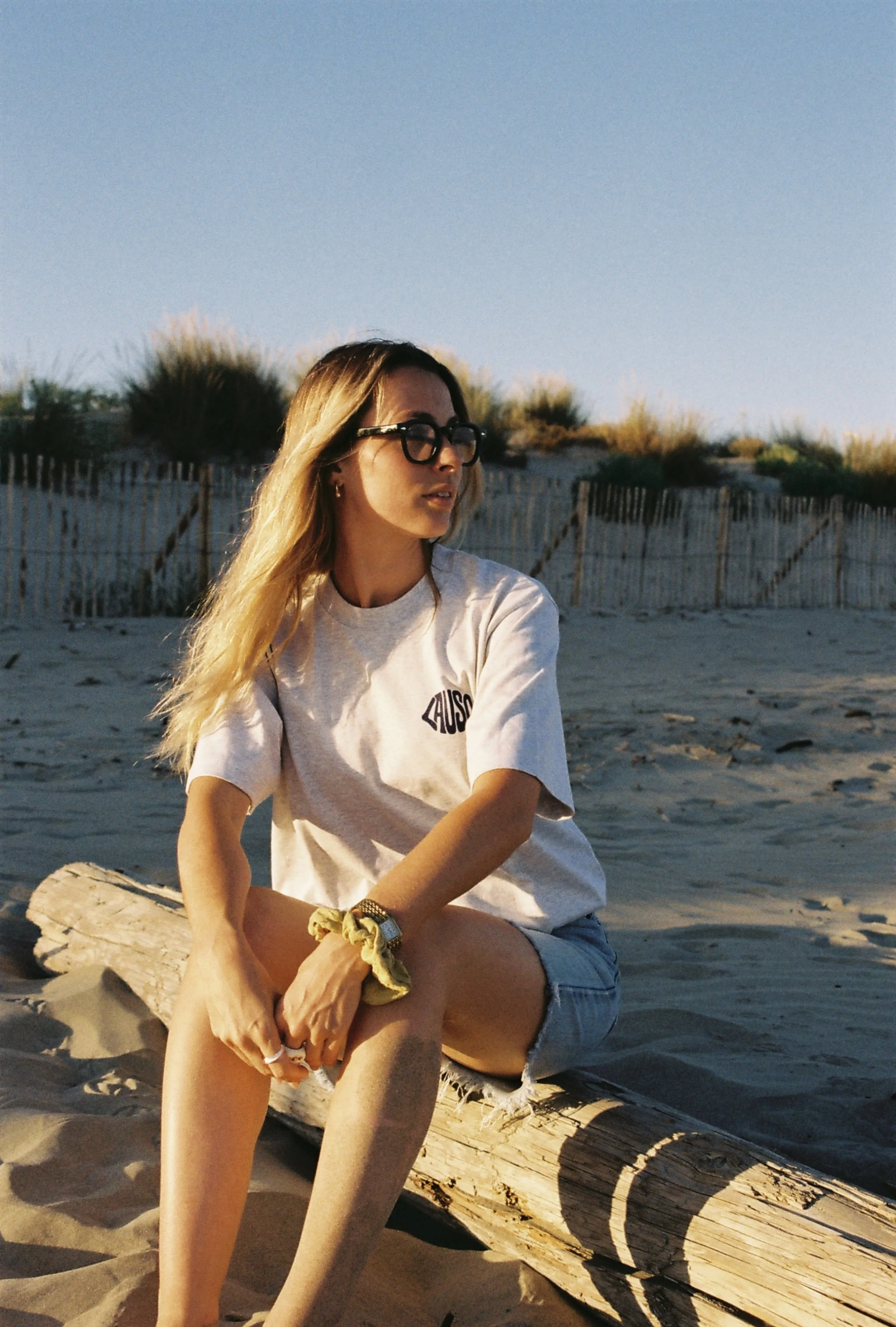 Jeune femme aux cheveux longs et blonds, portant des lunettes noires, une chemise beige avec le mot « AUSON » écrit dessus, assise sur un morceau de bois sur la plage, sous un ciel clair.